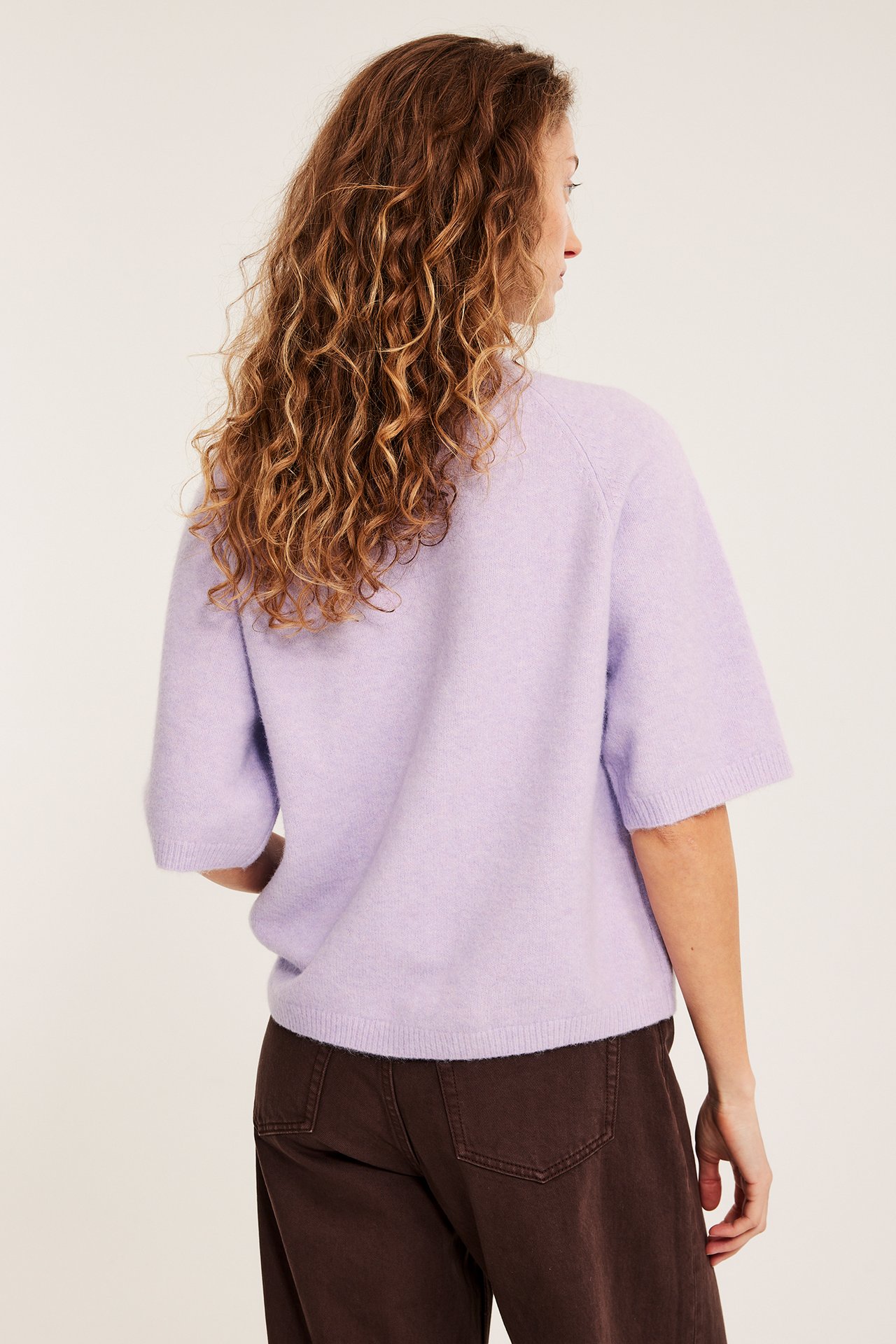Bakfra av en kvinne i en oversized lys lilla strikket T-skjorte over mørkebrune bukser.