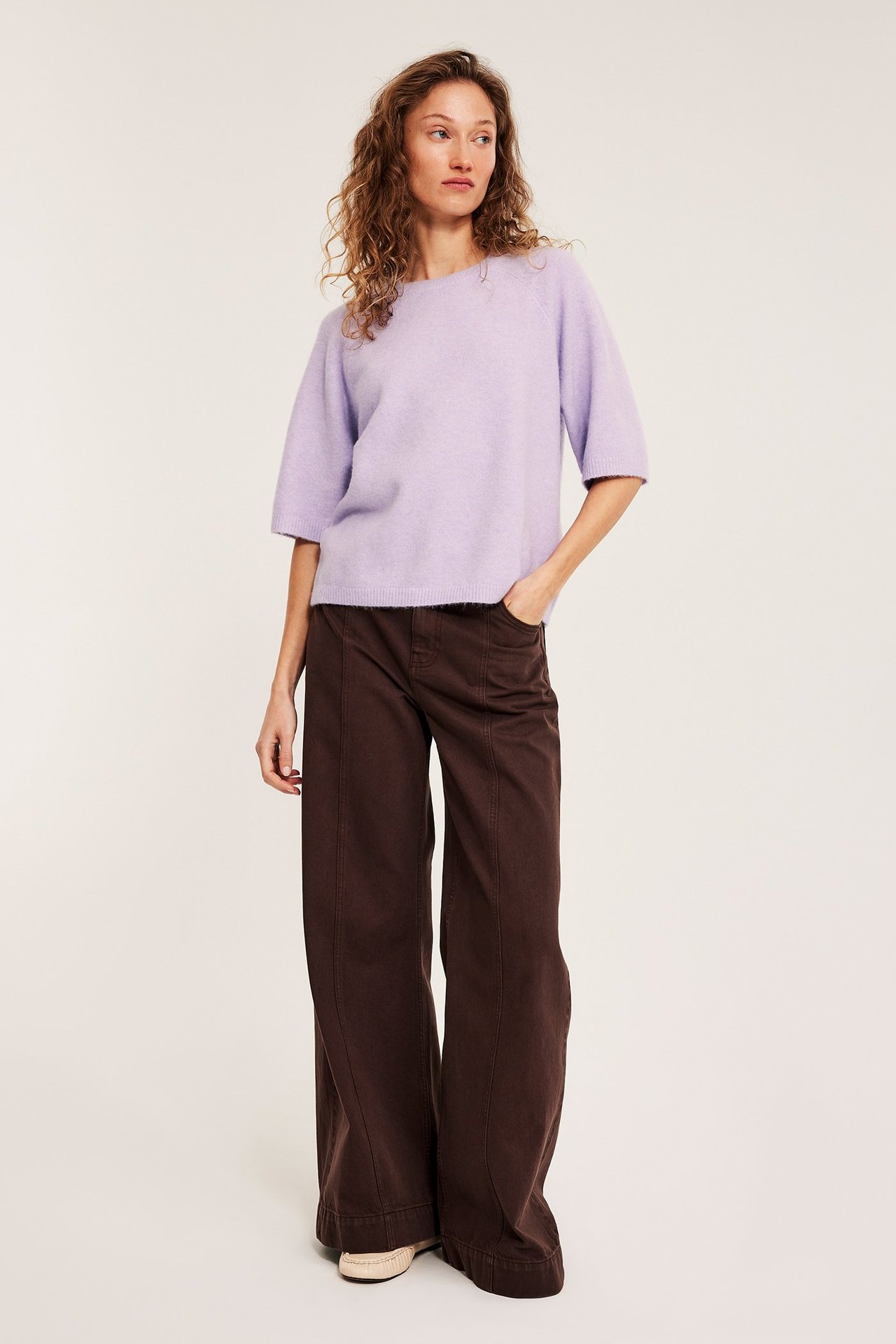 Edestäpäin kuvassa nainen, yllään oversized vaaleanlila neulepusero ja tummanruskeat leveälahkeiset housut.