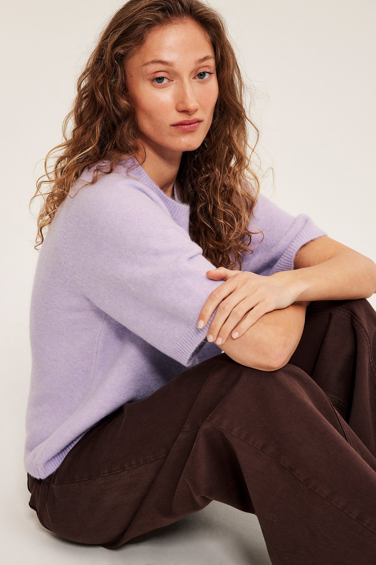 Lähikuva naisesta, jolla on ylisuuri vaalea lila neulottu t-paita ja ruskeat leveälahkeiset housut.