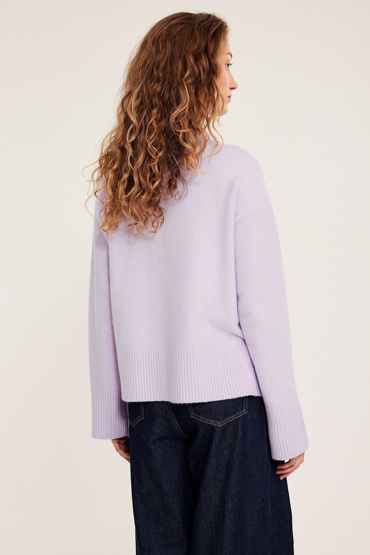 Widok z tyłu kobiety w jasnofioletowym dzianinowym swetrze i ciemnoniebieskich szerokich dżinsach.