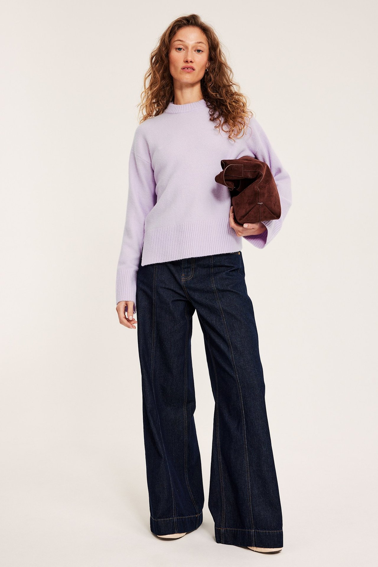 Framifrån: kvinna i ljuslila stickad pullover, mörkblå vida jeans, håller en brun kuvertväska.