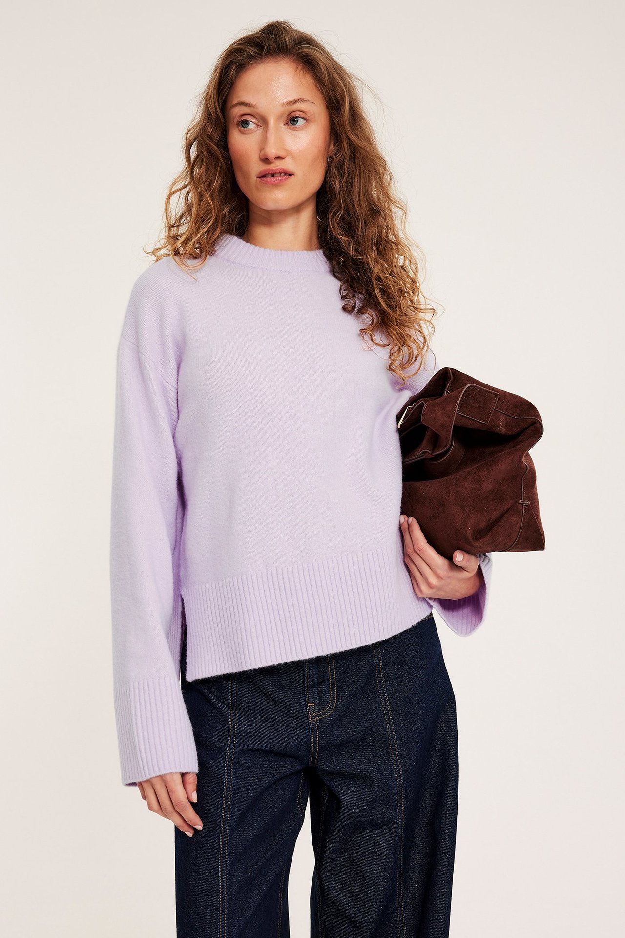 Kobieta ma na sobie jasnofioletowy dzianinowy sweter z okrągłym dekoltem, ciemnoniebieskie dżinsy, trzyma brązową zamszową torebkę.