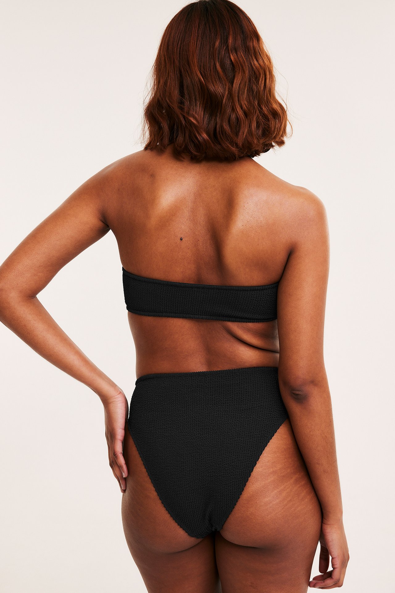 Nainen, takaa: mustat teksturoidut korkeavyötäröiset brazilian bikinialaosat ja bandeau-yläosa.