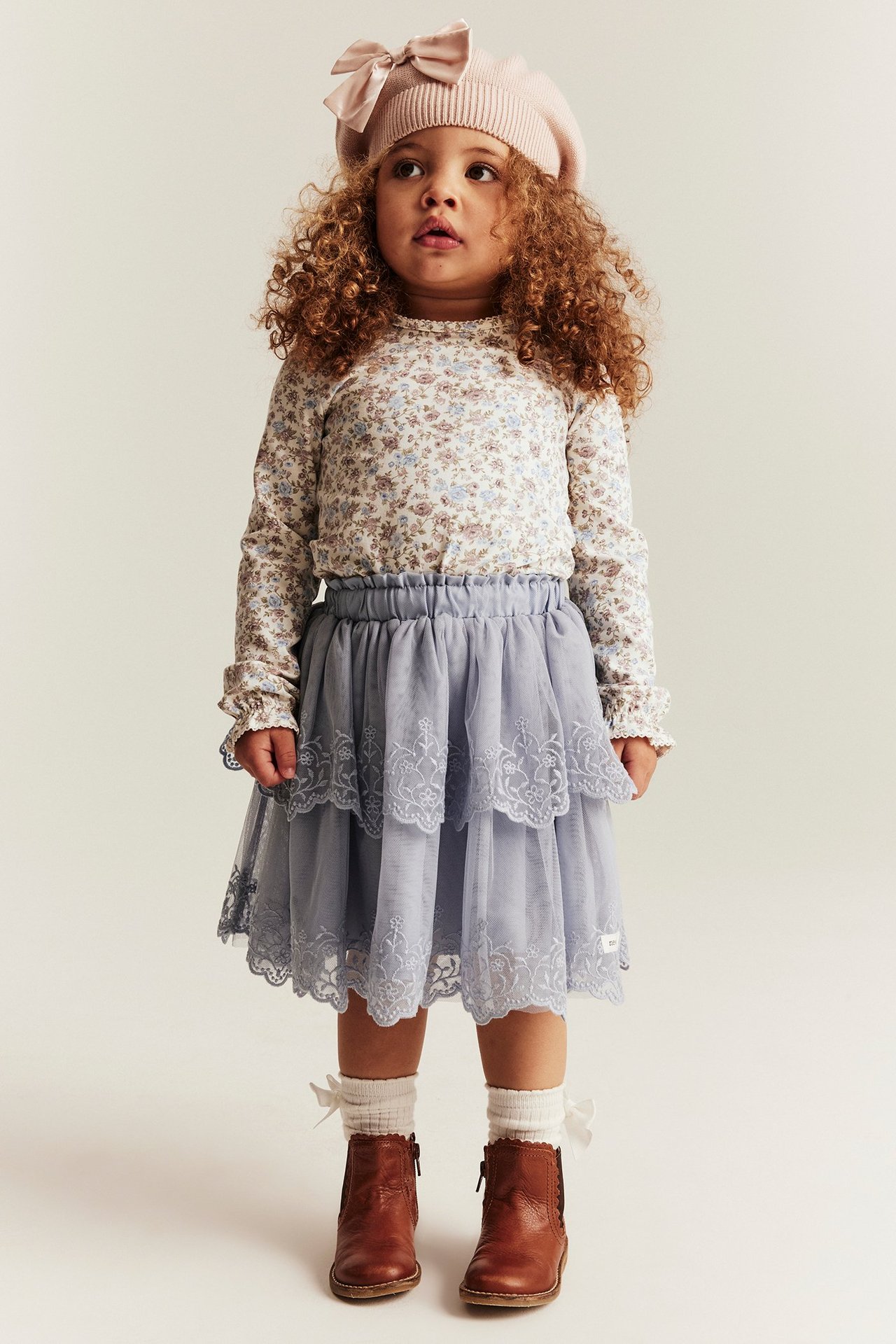 Forfra: barn i offwhite blomstret langermet topp, blå volangskjørt, rosa beret, hvite sokker, brune støvler.