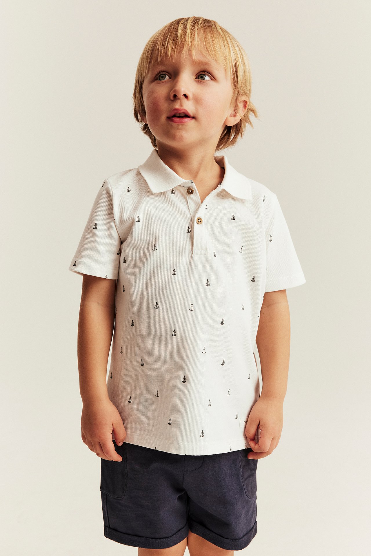 Framifrån, en pojke i offwhite bomullspiké med marint mönster och mörkblå shorts.