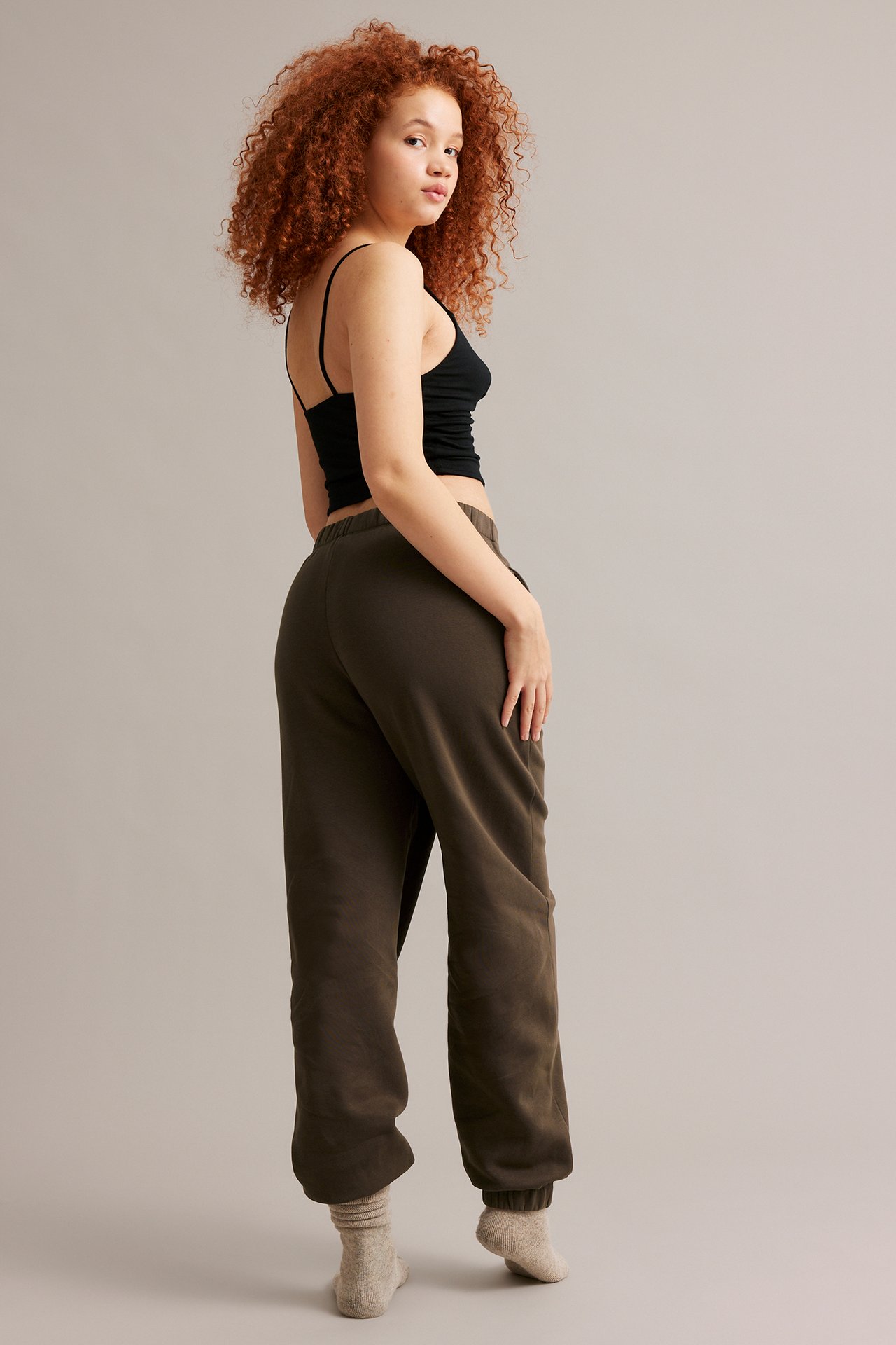 Takaapäin: nainen ruskeissa joggereissa sivutaskuilla, musta camisole ja vaaleanbeiget sukat.