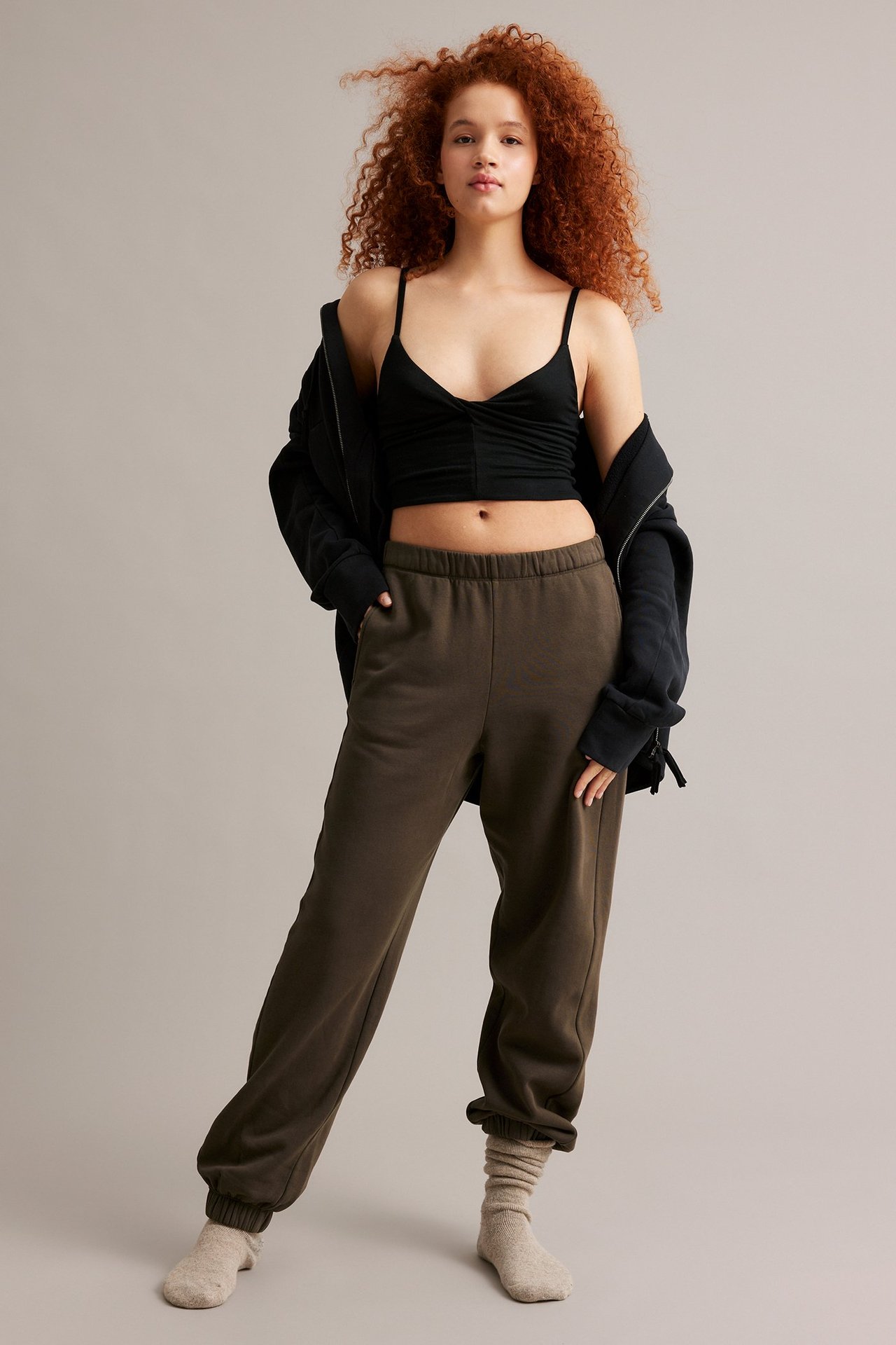 Kvinne i brune joggebukser, svart crop top, svart hettegenser og beige sokker.