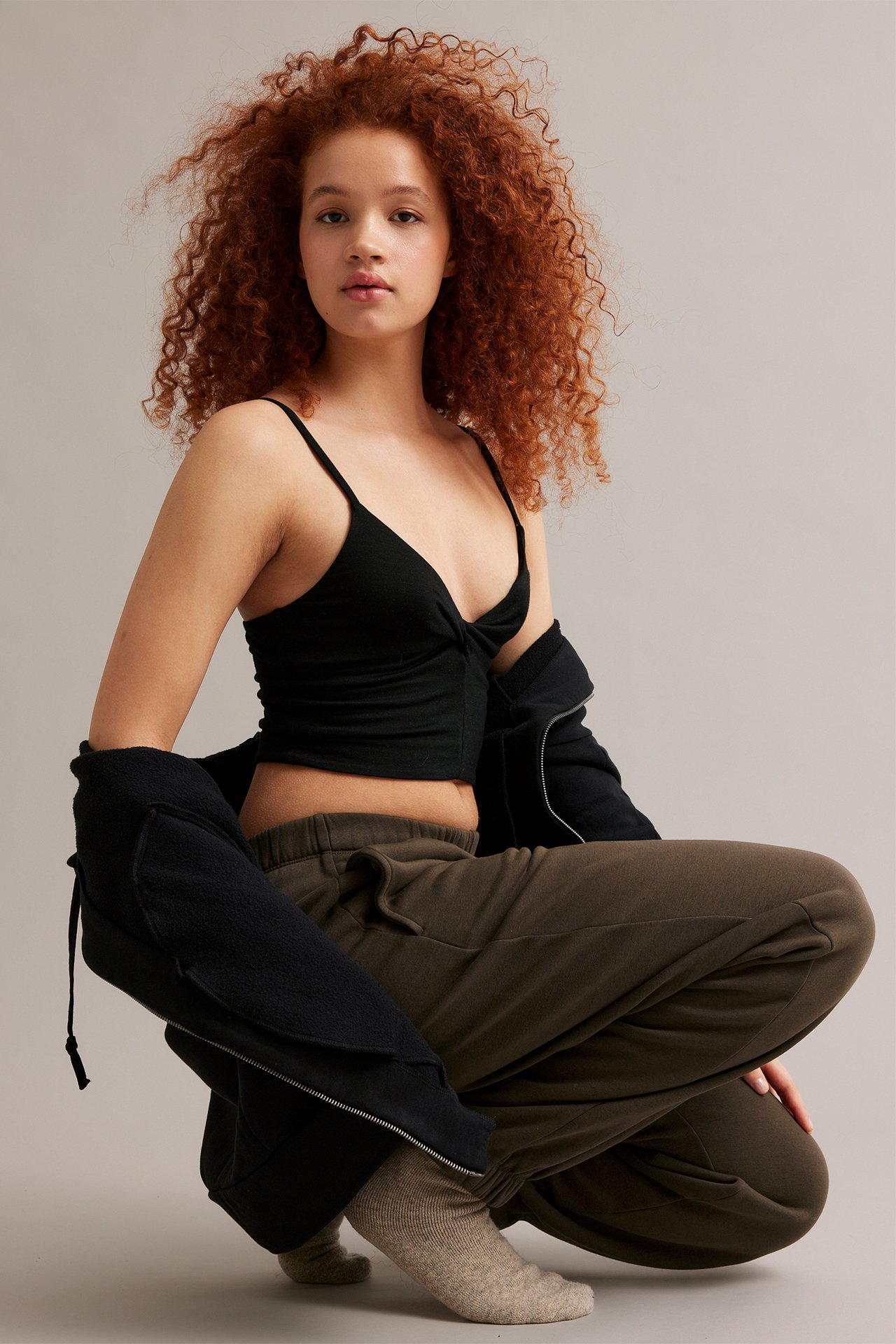 En kvinne forfra i brune joggere, en svart camisole og en svart hettegenser.