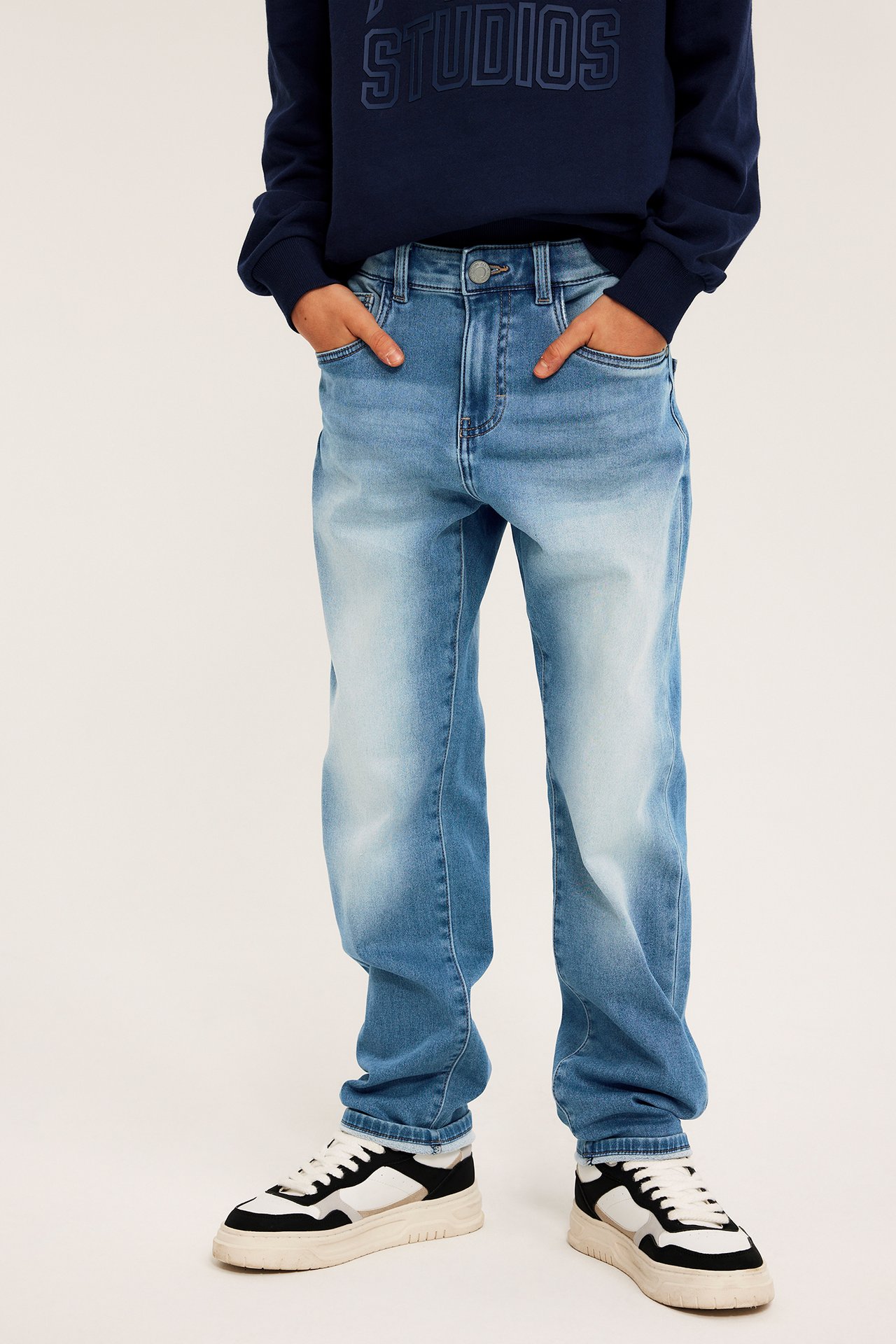 Forfra: Et barn har på seg lyseblå, avslappede jogger-jeans i denim, en marineblå genser og svarte/hvite joggesko.
