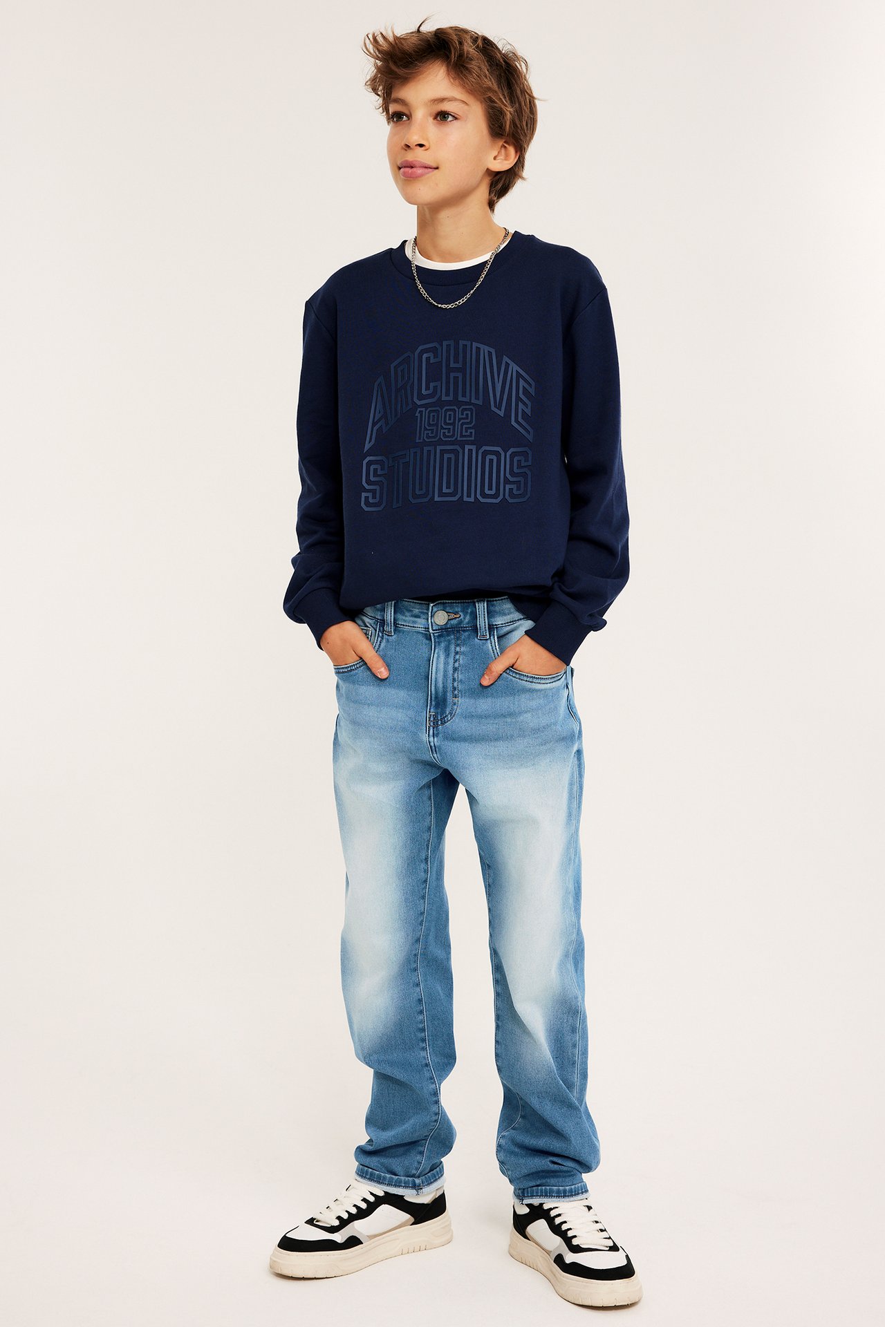 Barn i lyse relaxed joggerjeans, marineblå sweatshirt, svart-hvite sneakers, sølvkjede.