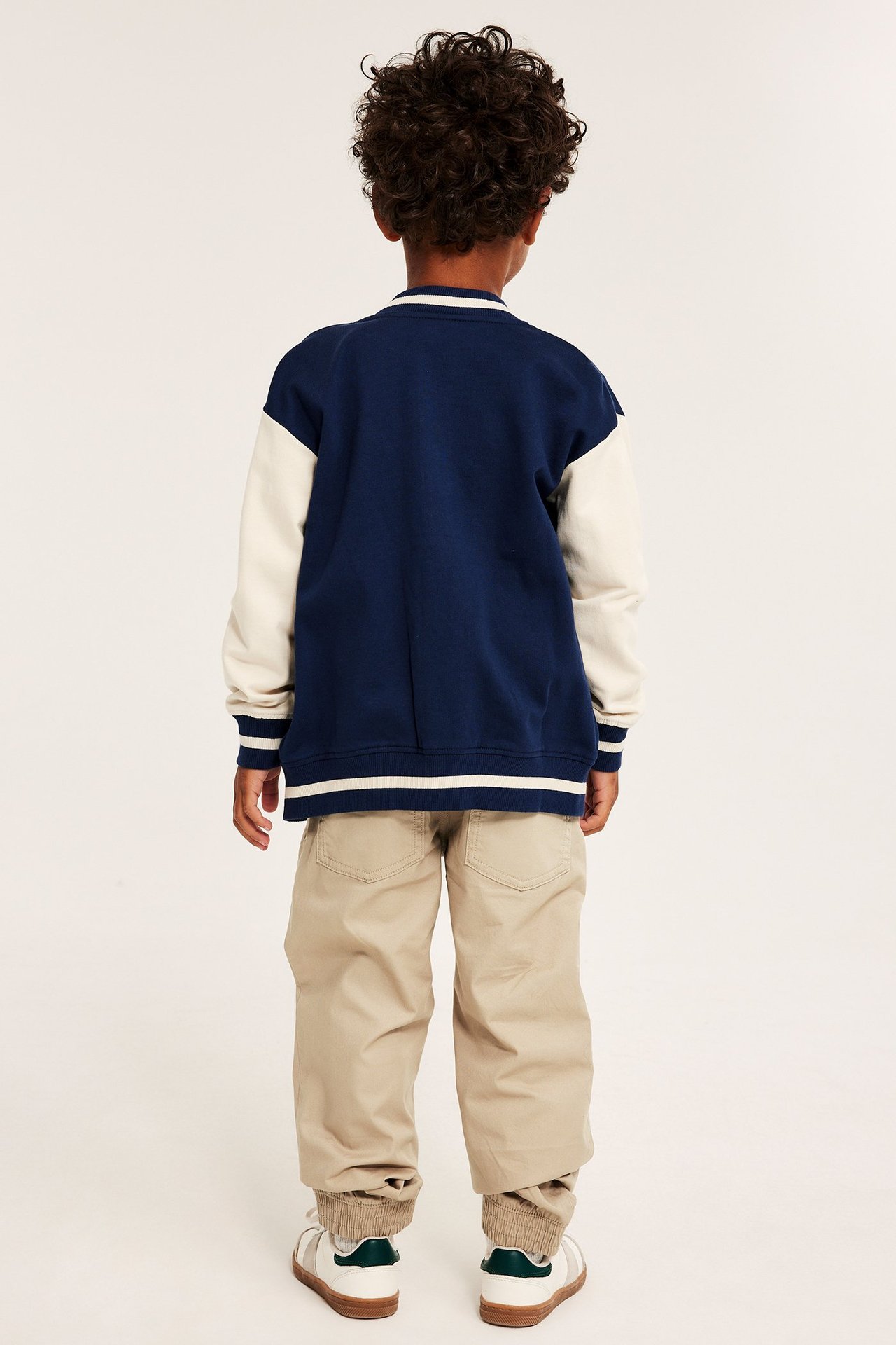 Barn bakifrån i beiga vävda bomullsbyxor med resår, en marinblå/krämfärgad varsityjacka och vita sneakers.