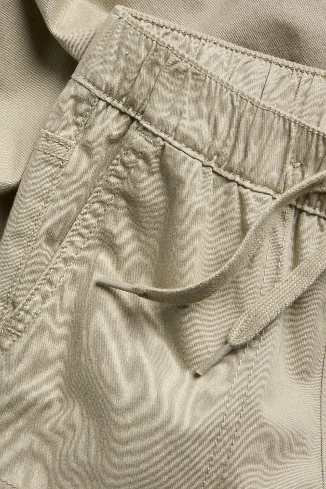 Zbliżenie beżowych tkanych spodni bawełnianych z elastycznym pasem, sznurkiem i kieszenią boczną.
