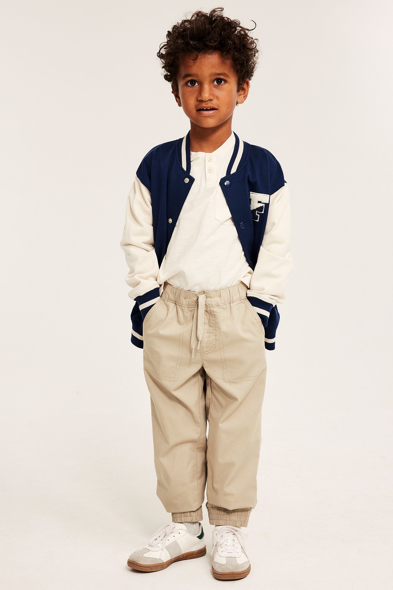 Frontvy av barn i beiga bomullsbyxor med dragsko, resårmuddar, vit tröja och collegejacka.