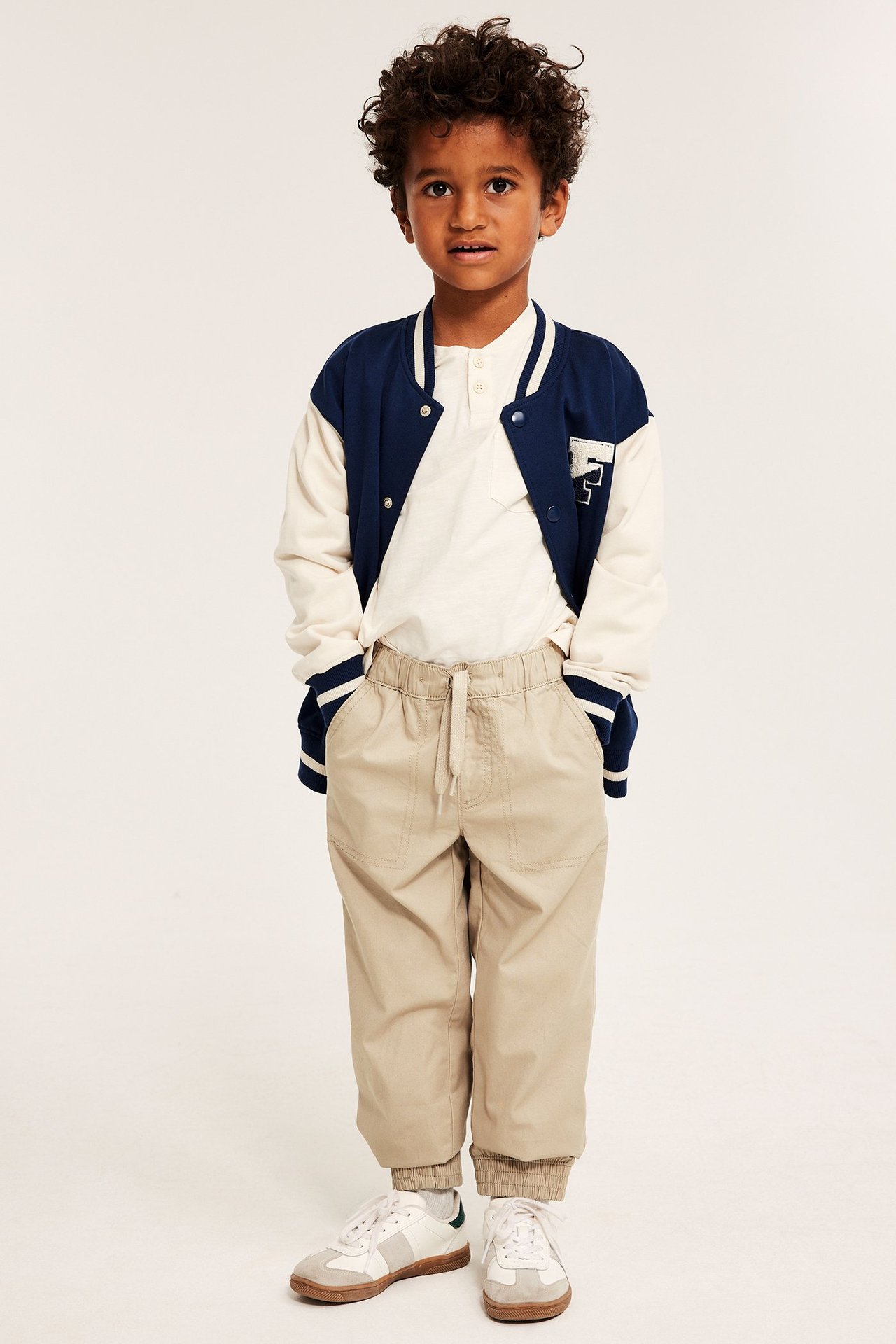 Edestäpäin lapsi beigessä puuvillahousuissa, joissa kiristysnyöri ja joustavat suut, valkoinen paita ja takki.