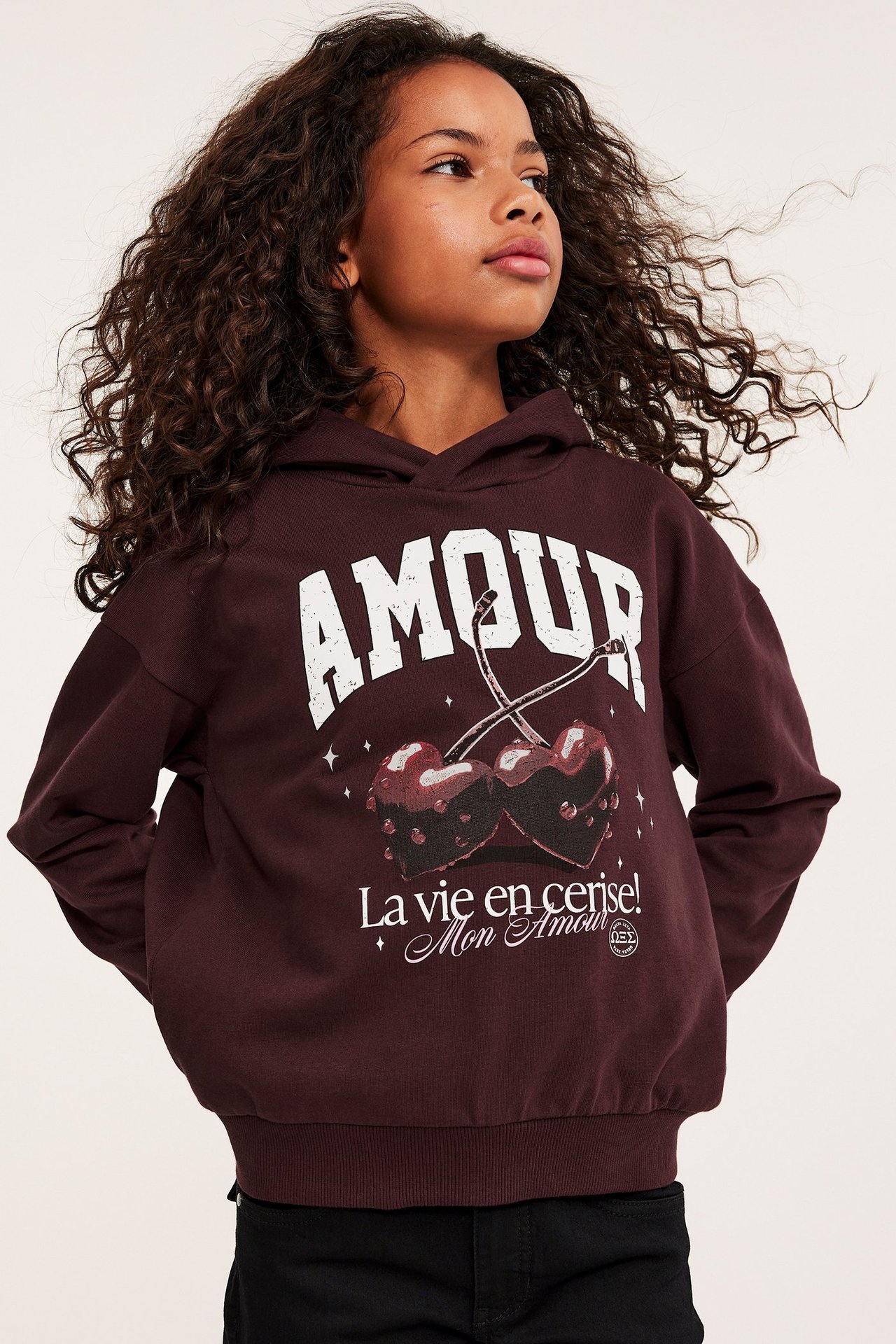 Edestä kuvattu lapsi tummanpunaisessa puuvillahupparissa, jossa valkoinen 'AMOUR' ja kirsikkaprintti.