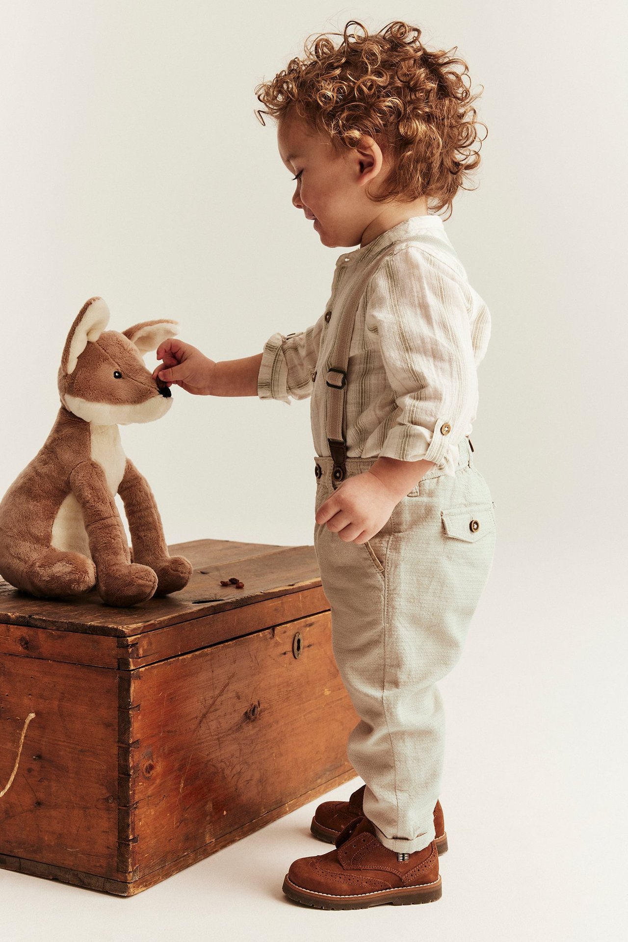 Sivukuva vauvasta beigeissä puuvillahousuissa henkseleillä, raidallisessa paidassa ja ruskeissa kengissä.