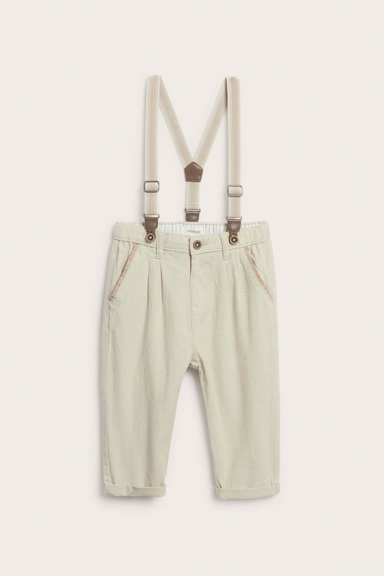 Z przodu: beżowe tkane spodnie dziecięce z regulowanymi szelkami i podwiniętymi nogawkami.
