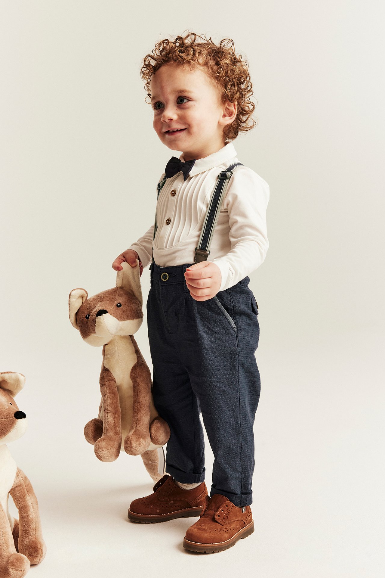 Edestä: Lapsi tummansinisissä kudotuissa henkselihousuissa, valkoisessa laskostetussa paidassa, mustassa rusetissa, pitäen kettulelua.