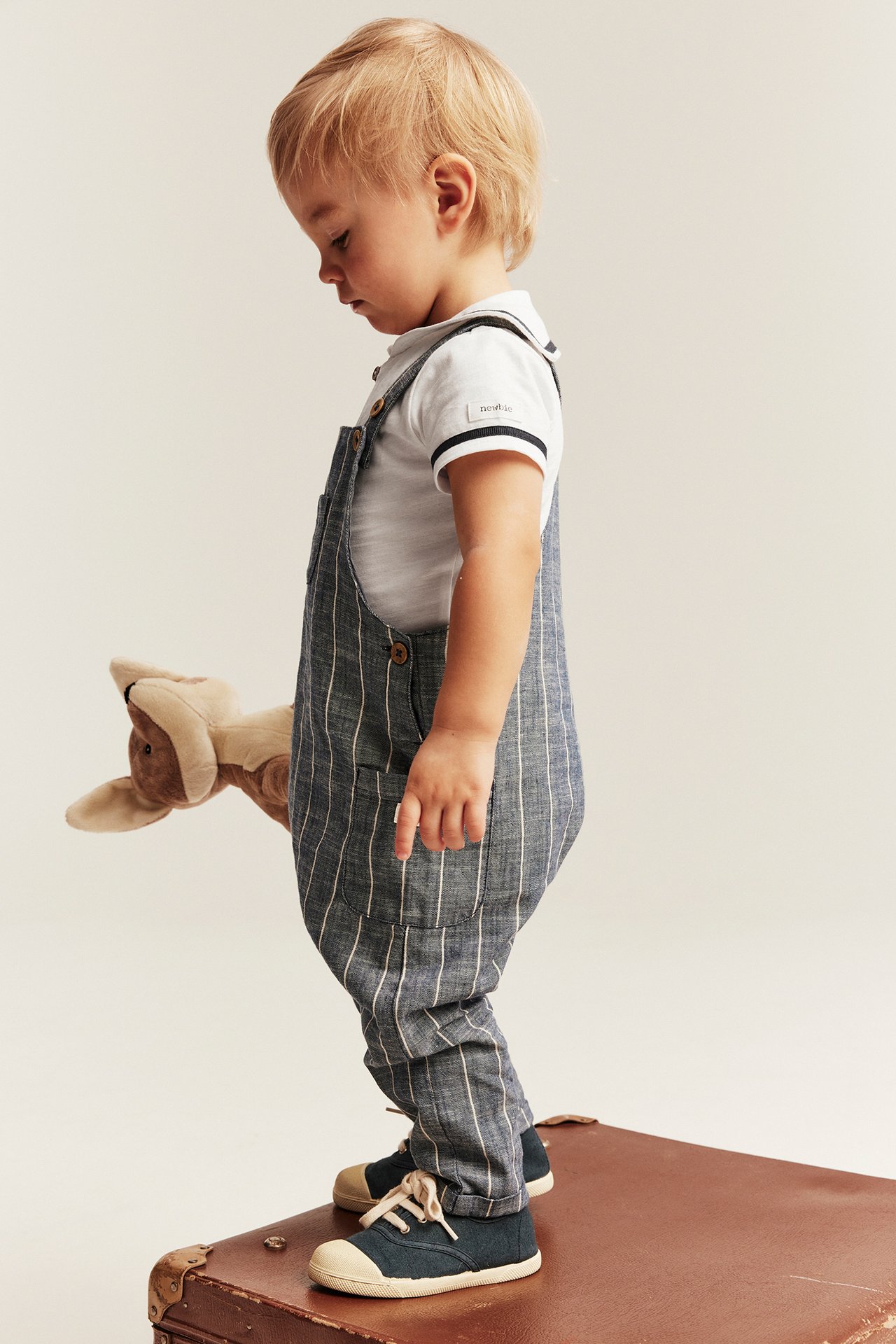 Sidevisning av baby i offwhite sjømannsbody, blåstripete overall, mørkeblå tøysko, holder en kenguruleke.