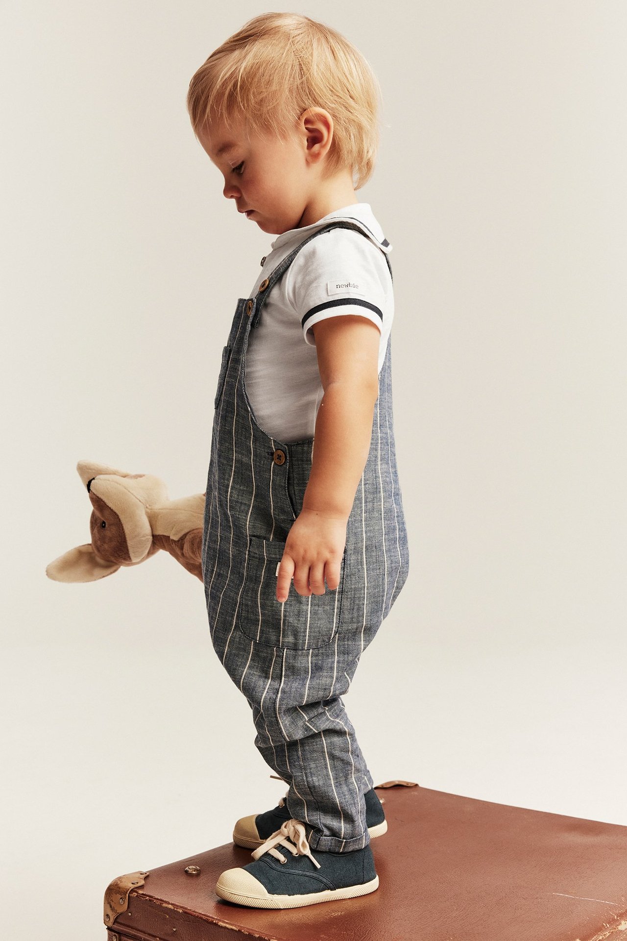 Sidevisning av baby i offwhite sjømannsbody, blåstripete overall, mørkeblå tøysko, holder en kenguruleke.