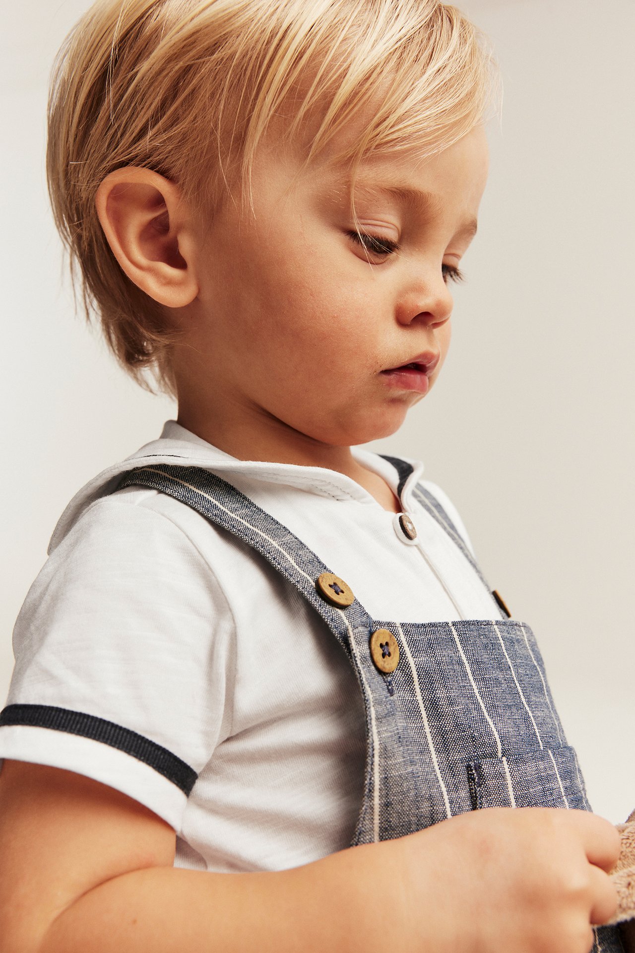 Nærbilde i sideprofil av en baby i en offwhite kortermet matrosbody under blåstripete snekkerbukse med treknapper.