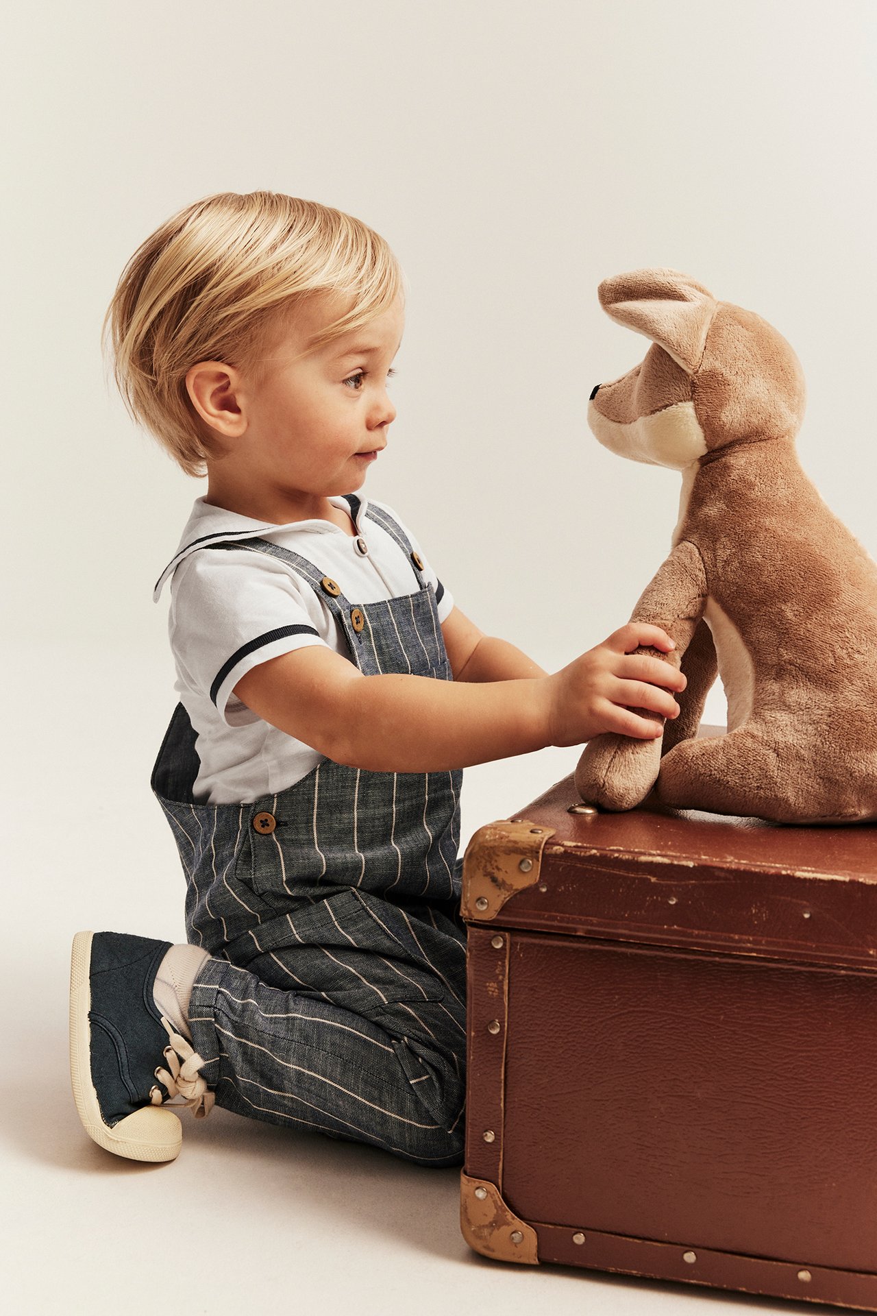 Sidevisning: baby i offwhite matrosbody, stripete snekkerbukse, blå sko, holder et brunt kosedyr.