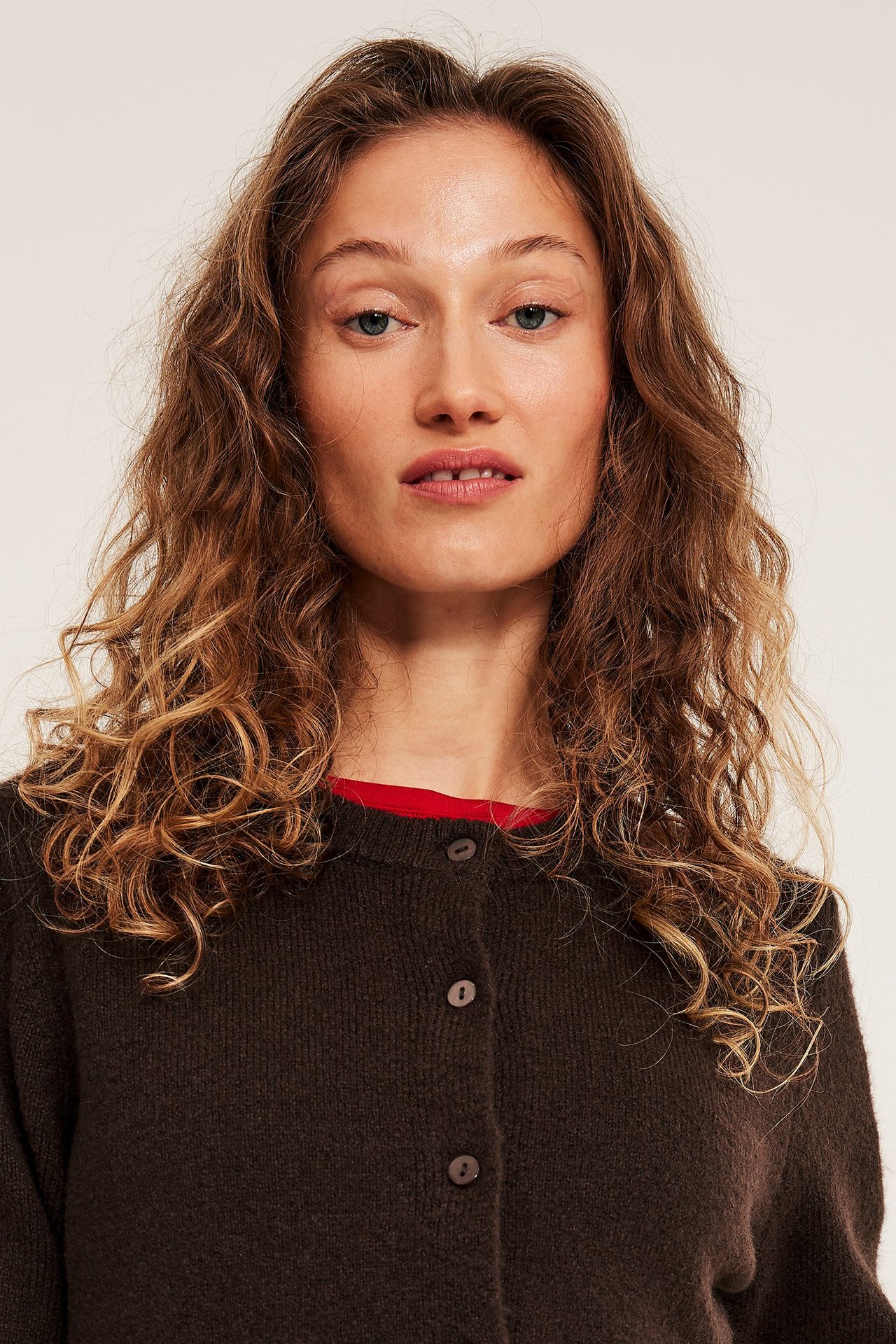 Lähikuva naisesta tummanruskeassa neulotussa lyhythihaisessa napitetussa neuletakissa, jonka alla punainen toppi.