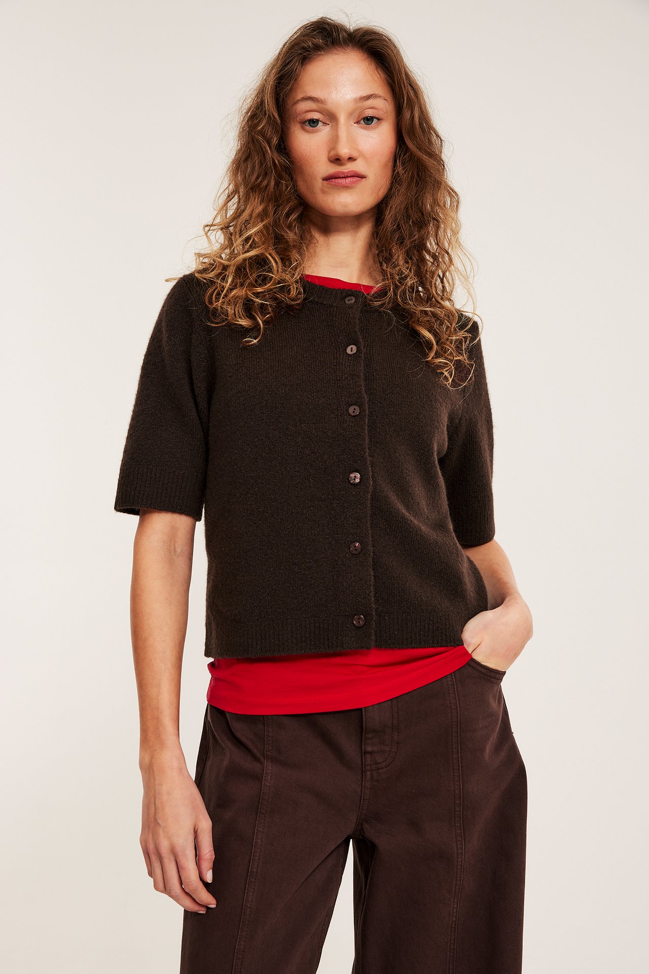 Nainen edestäpäin yllään tummanruskea lyhythihainen nappineuletakki, punainen toppi ja tummanruskeat leveälahkeiset housut.