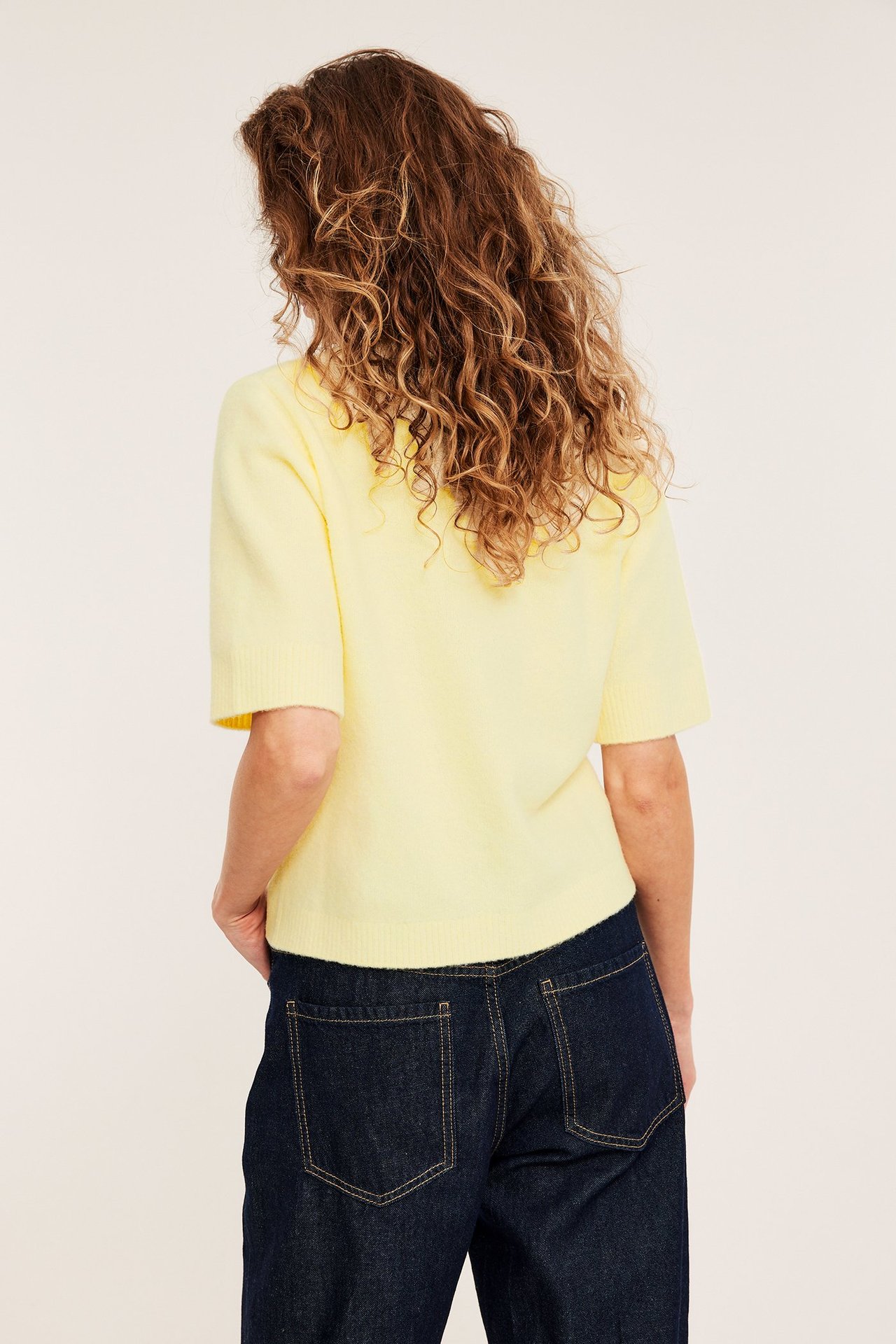 Naisen takaapäin, yllään vaaleankeltainen lyhythihainen neulepusero ja tummansiniset farkut.