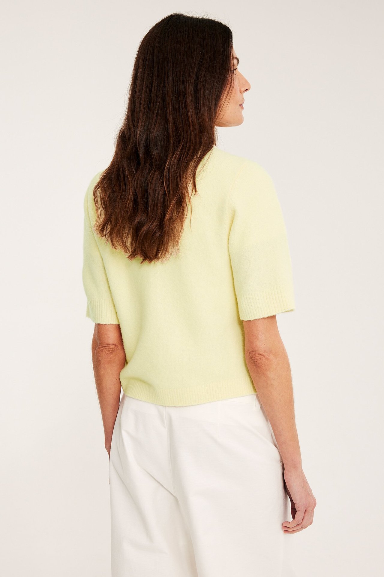 Widok z tyłu kobiety w jasnożółtym swetrze z krótkim rękawem i białych szerokich spodniach.