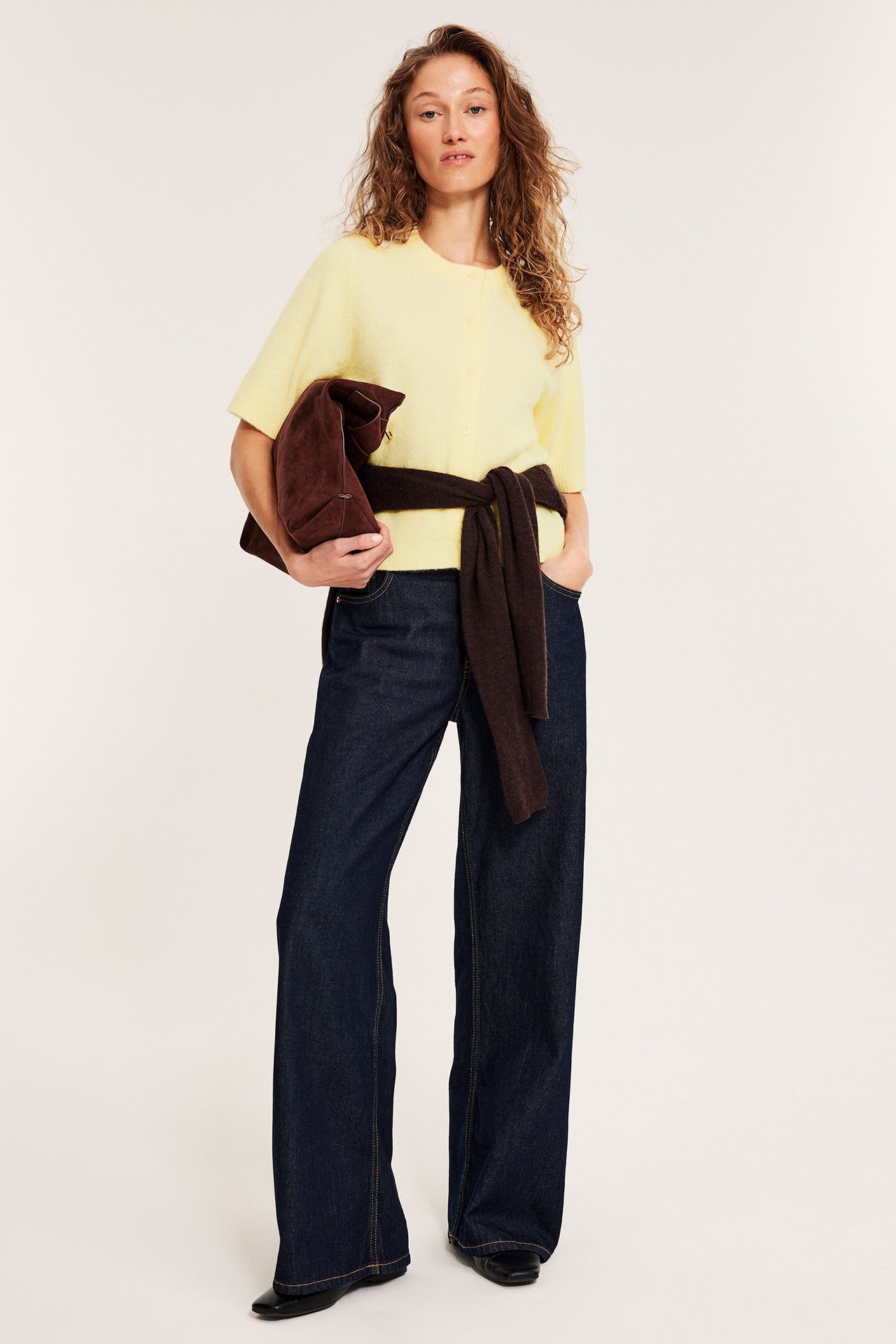 Nainen edestä, vaaleankeltainen lyhythihainen neuletakki, tummansiniset farkut, ruskea laukku.