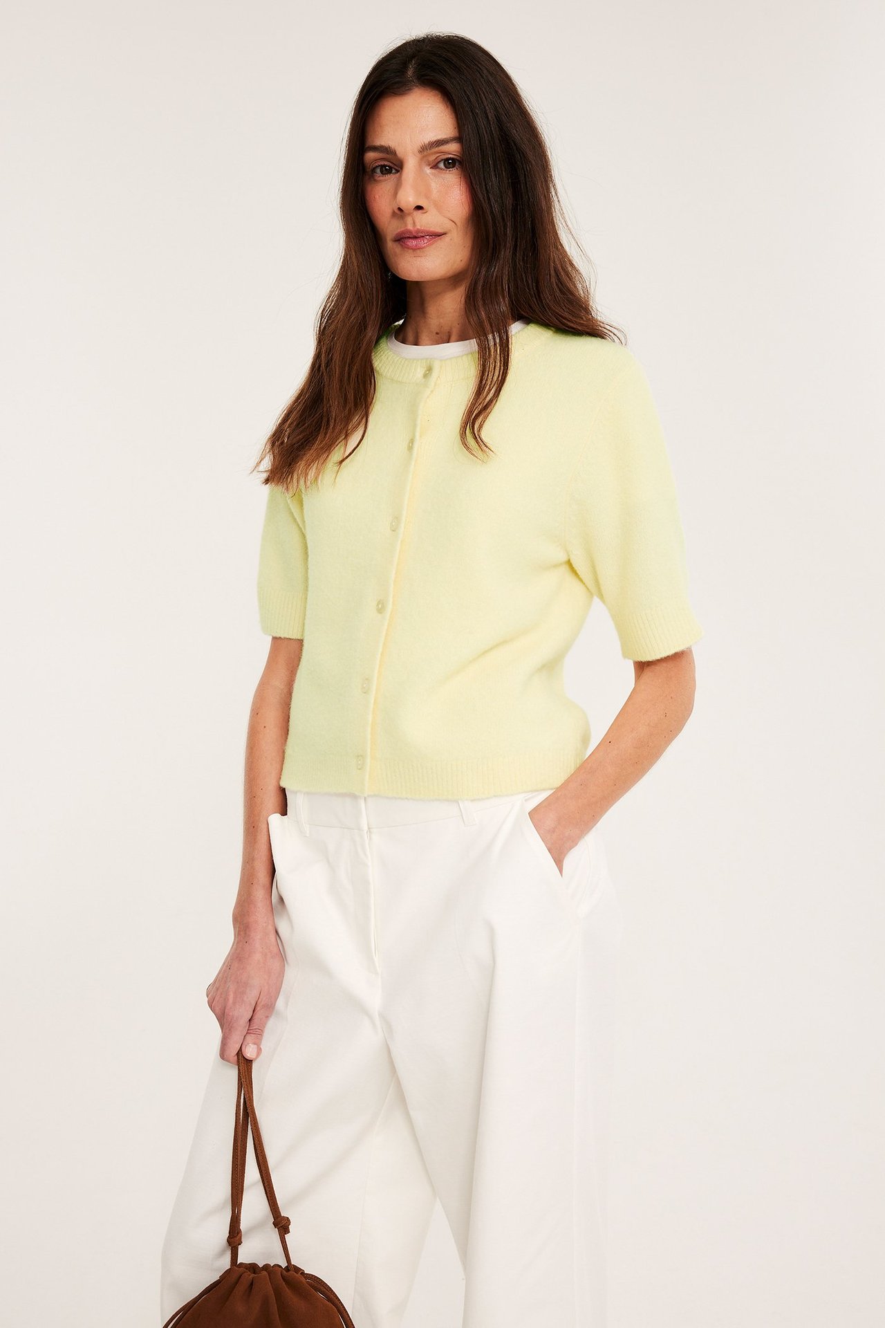 Widok z przodu: kobieta w jasnożółtym swetrze rozpinanym, białym topie, białych spodniach, trzyma brązową torbę.