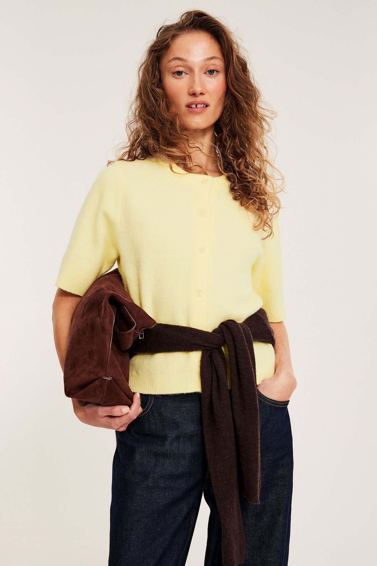 Kvinna bär en ljusgul kortärmad stickad kofta, mörka jeans, håller brun väska, med brun stickad tröja knuten i midjan.