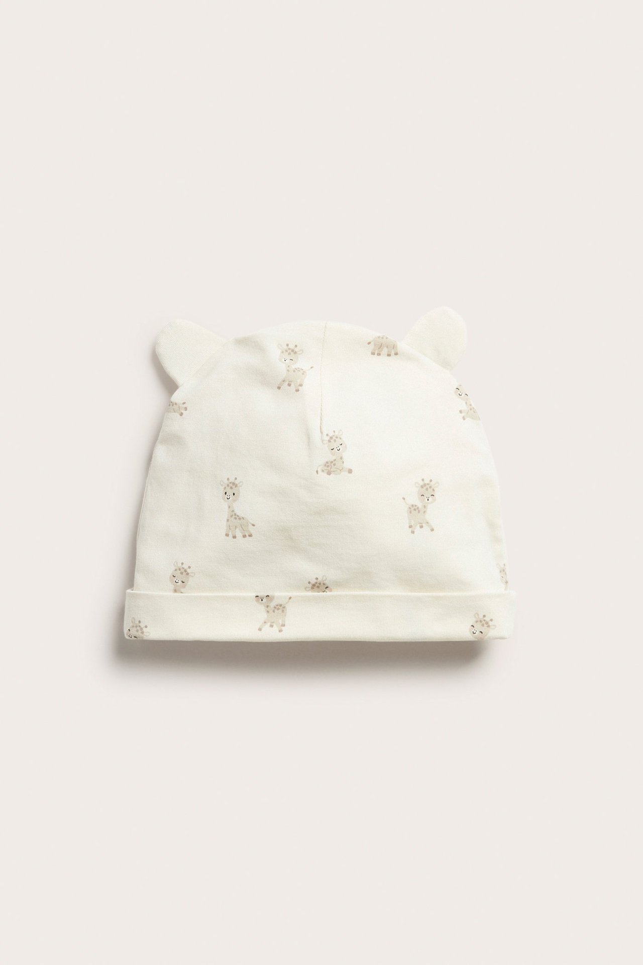 Vaaleabeige jersey-vauvanpipon etunäkymä, jossa on korvat, käännetty reuna ja kirahvikuosi.