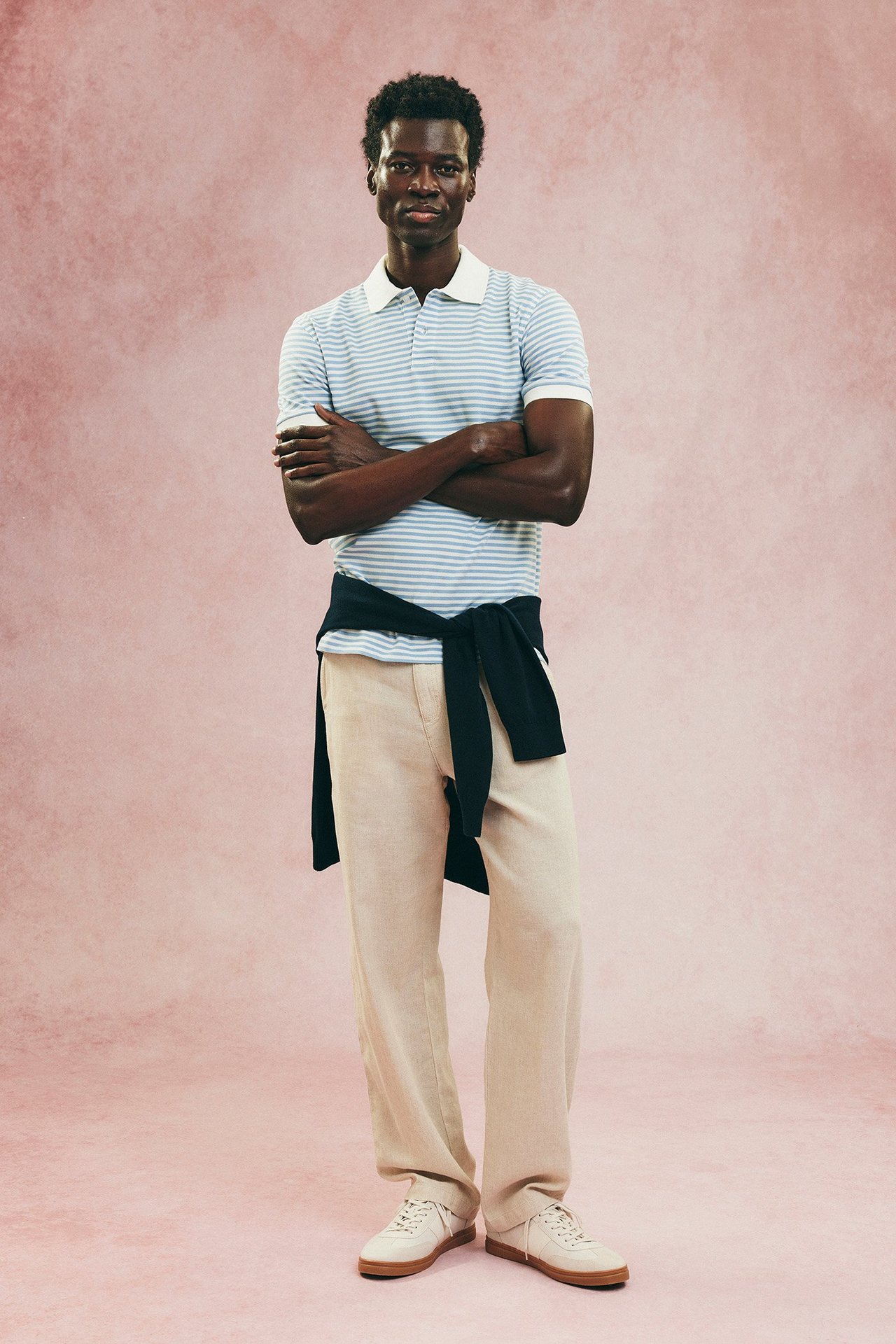 Mies yllään beiget pellavasekoitehousut, vaaleansininen raidallinen pikeepaita, tumma pusero vyötäröllä, valkoiset tennarit.