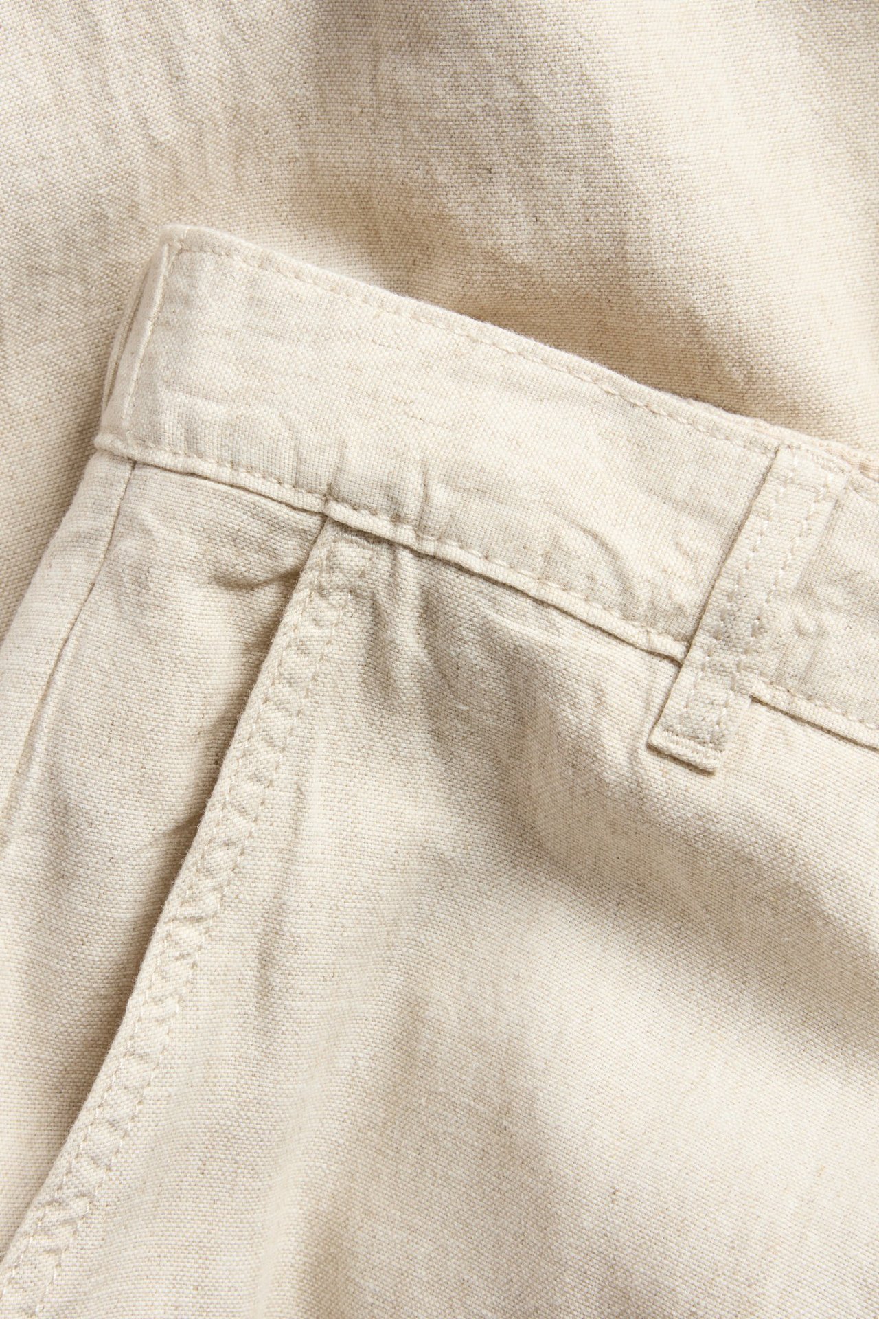 Zbliżenie beżowych spodni z mieszanki lnu o regularnym kroju, widać pas, szlufkę i otwór kieszeni.
