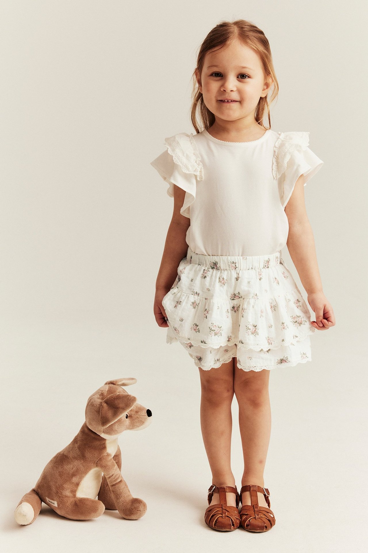 Widok z przodu: dziecko w kremowych kwiatowych spodenkach-spódnicy, kremowym topie i brązowych sandałach.