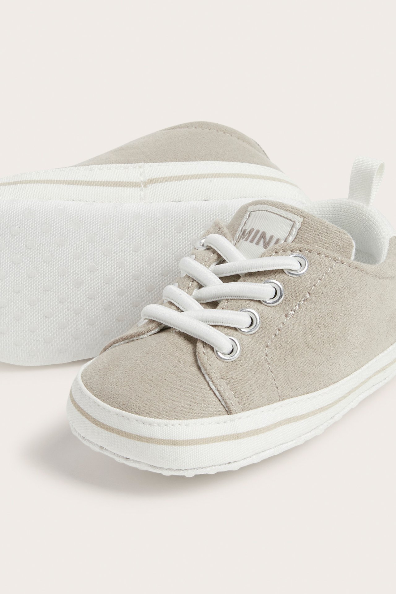 Nærbilde av beige sneakers i semsket imitasjon for baby med hvite lisser, antiskli-såle og brodert tunge.