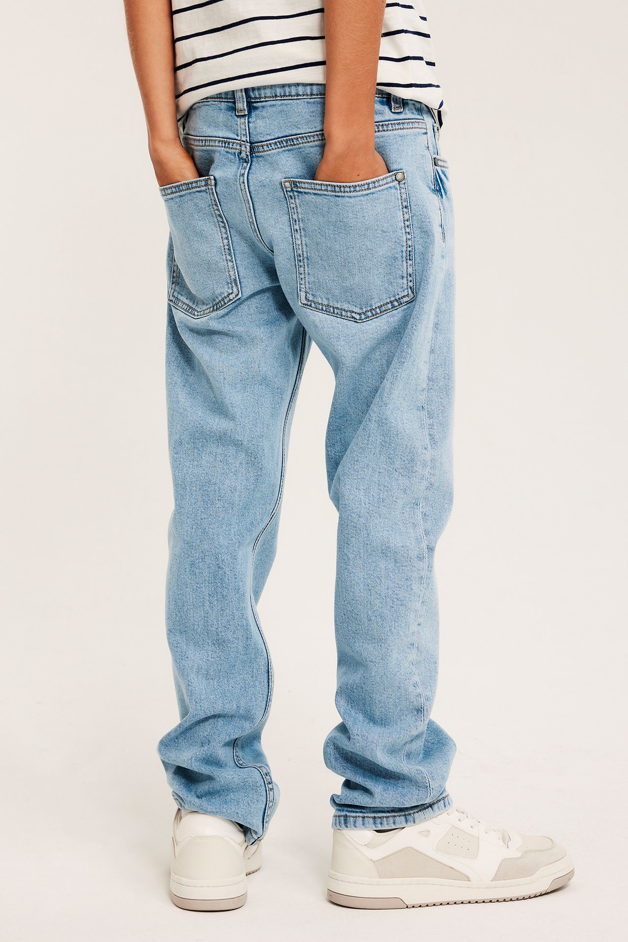 Widok z tyłu dziecka w jasnoniebieskich jeansach, ręce w kieszeniach, pasiasta koszulka i sneakersy.