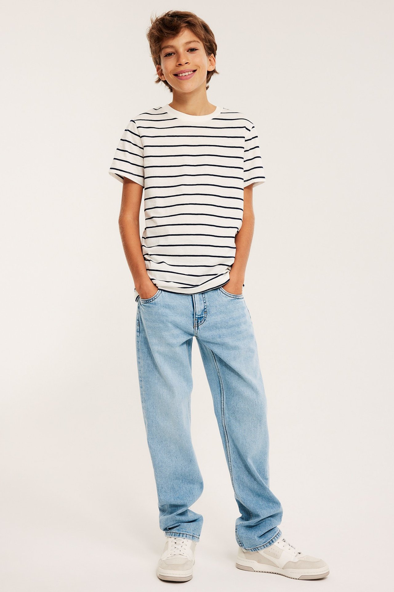 Edestä kuvattu hymyilevä lapsi yllään vaaleansiniset suorat farkut, valkoinen raidallinen t-paita ja valkoiset tennarit.
