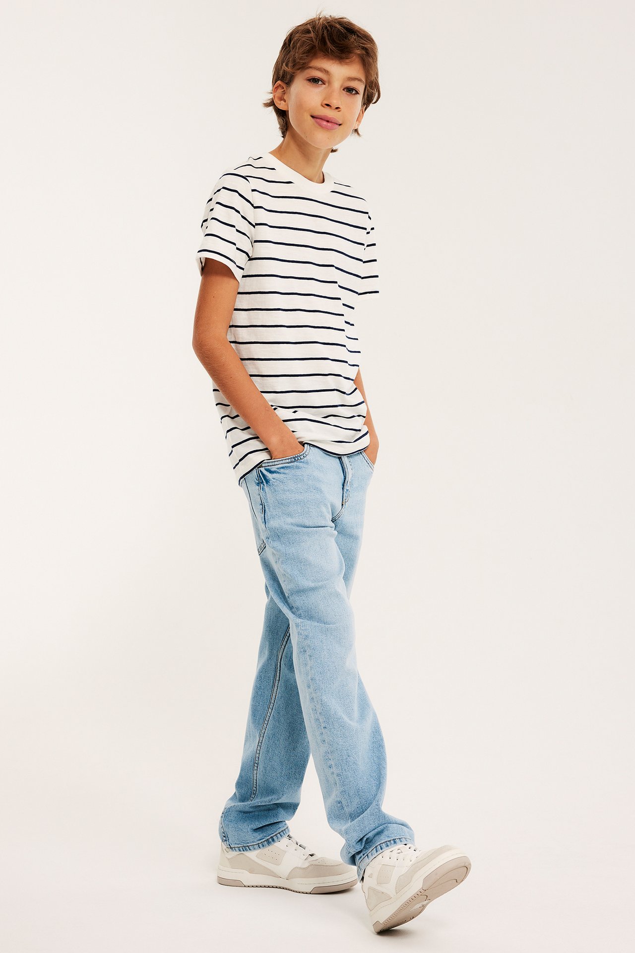 Z przodu: Dziecko w jasnoniebieskich prostych jeansach, pasiastym t-shircie i jasnych butach.