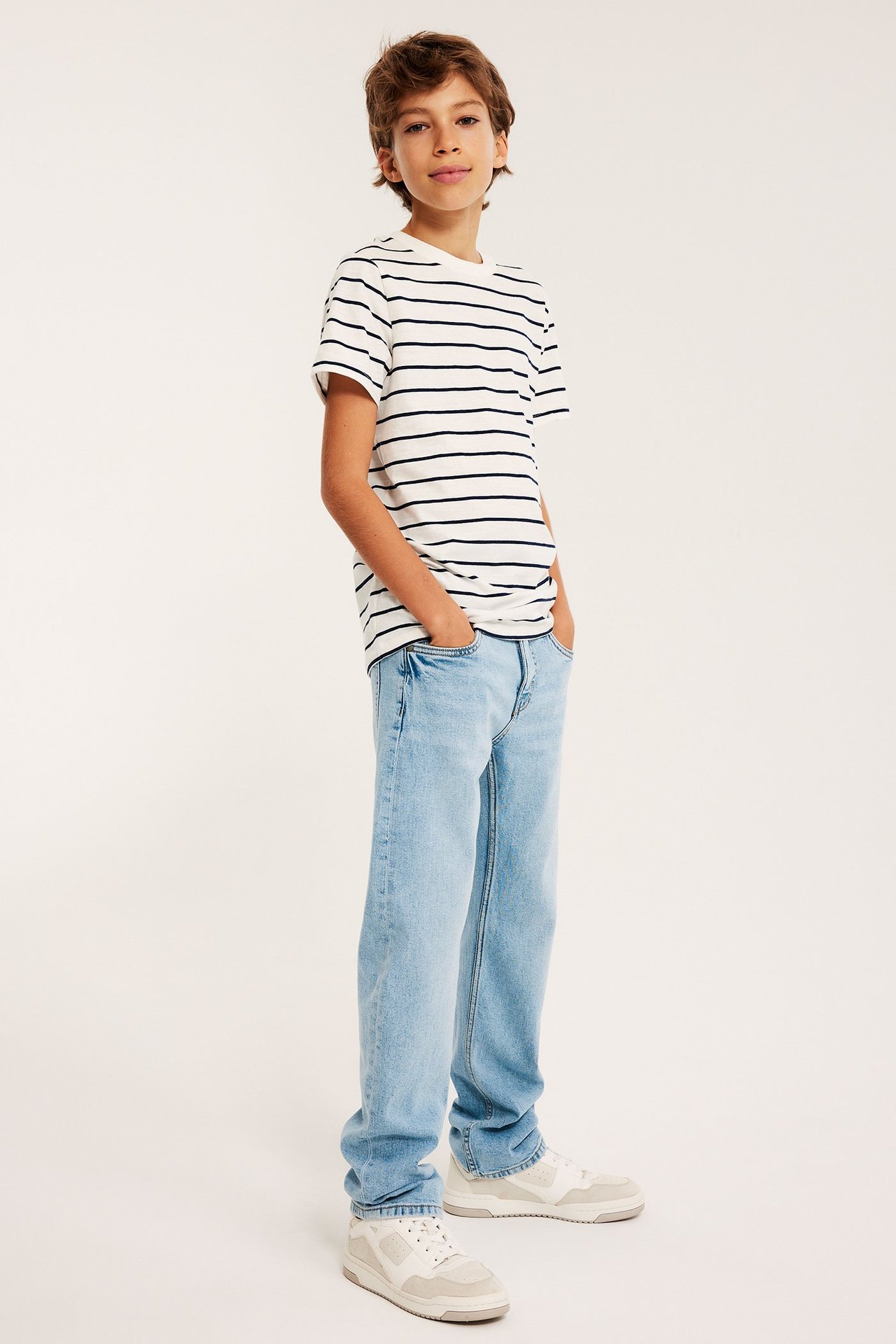 Widok z przodu: dziecko w jasnoniebieskich prostych jeansach, białym pasiastym t-shircie, jasnych sneakersach.