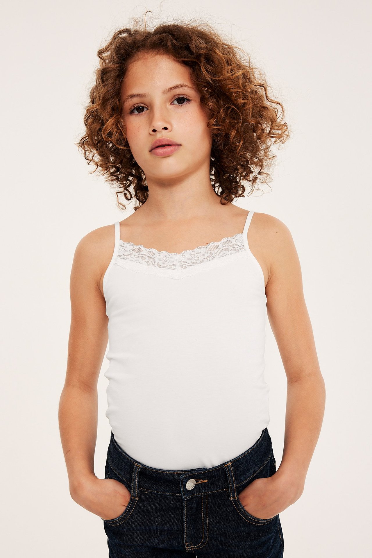 Framifrån: ett barn i offwhite ribbat linne med vit spetskant och mörkblå jeans.