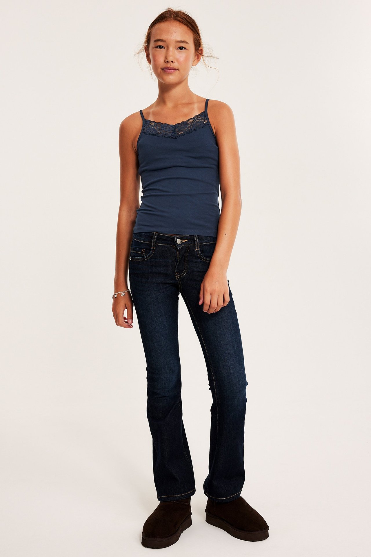 Frontvy av en tjej som bär ett blått ribbat linne med spetskant, mörkblå jeans och mörkbruna stövlar.