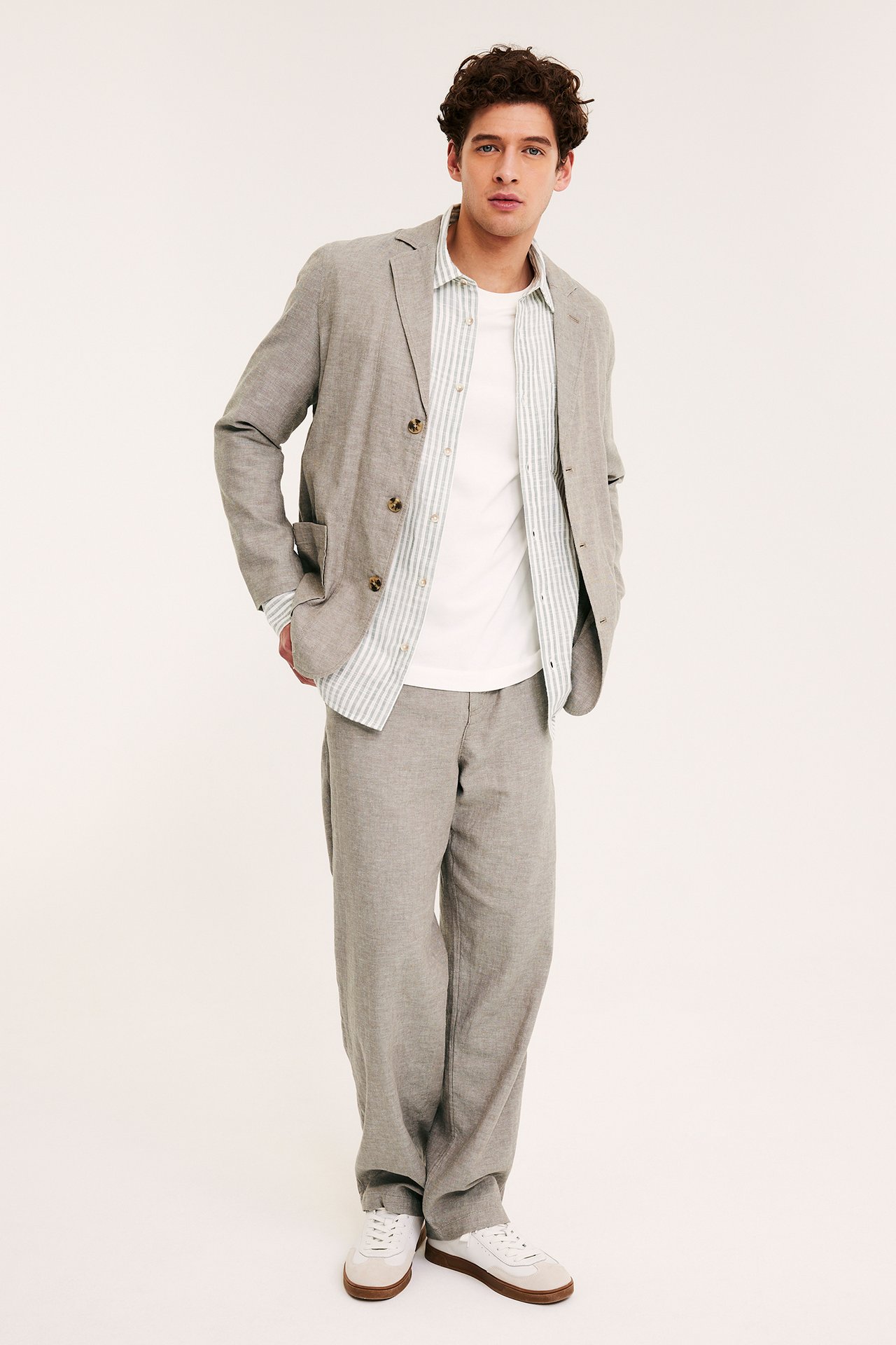 Mężczyzna w jasnobrązowej marynarce z mieszanki lnu, koszuli w paski, pasujących spodniach i białych trampkach.