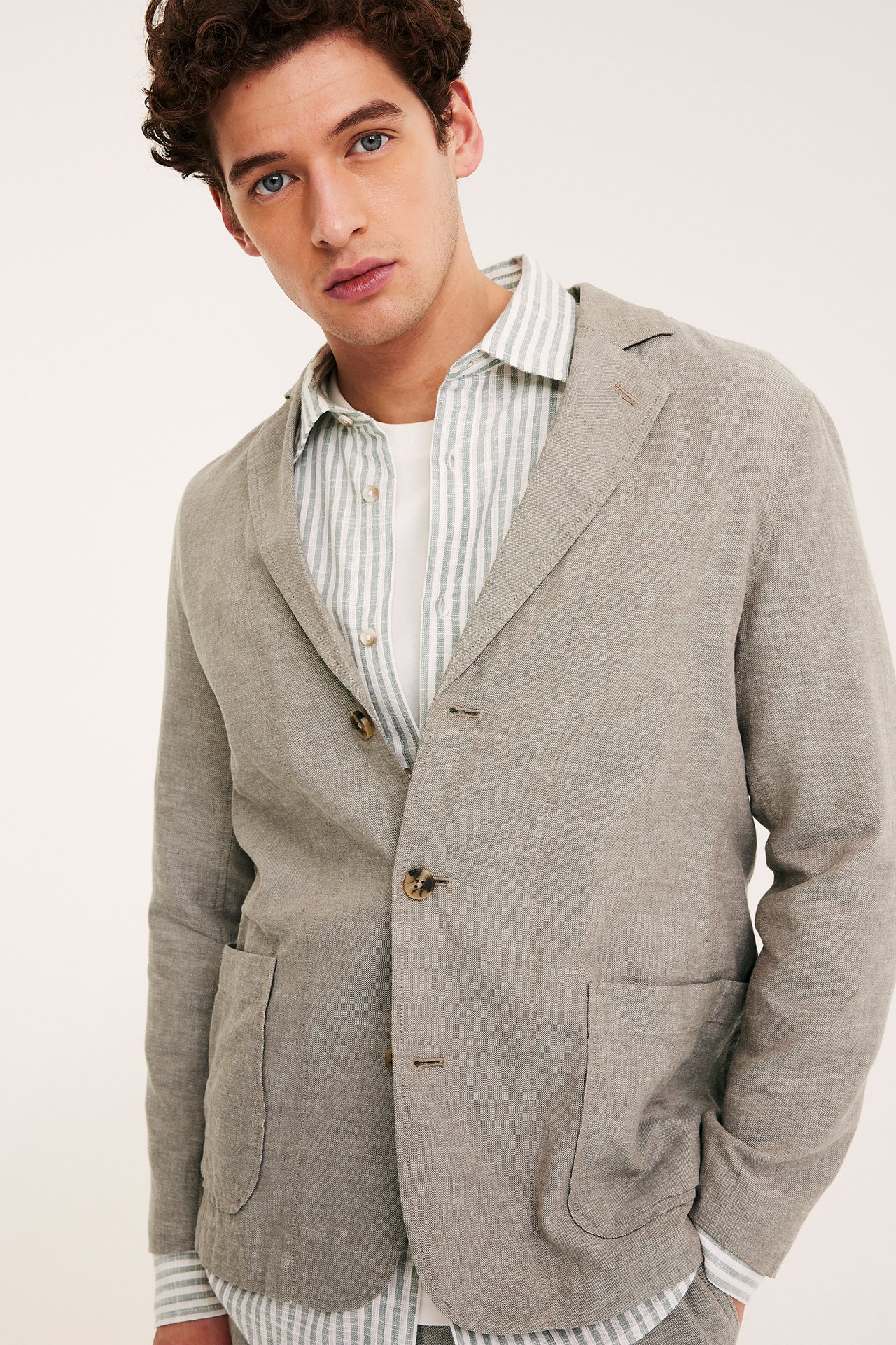 Widok z przodu: mężczyzna w otwartej brązowej lnianej marynarce, na zielonej koszuli w paski i białym t-shircie.