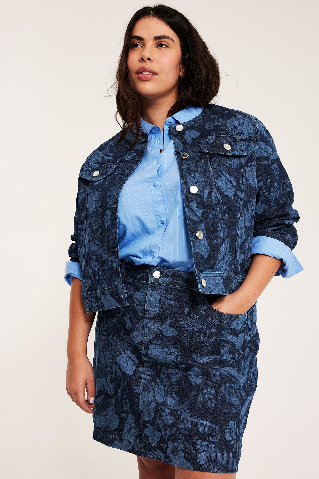 Edestä: Nainen sinisessä kukkakuosisessa farkkuhameessa, vastaavassa farkkutakissa ja vaaleansinisessä raidallisessa paidassa.