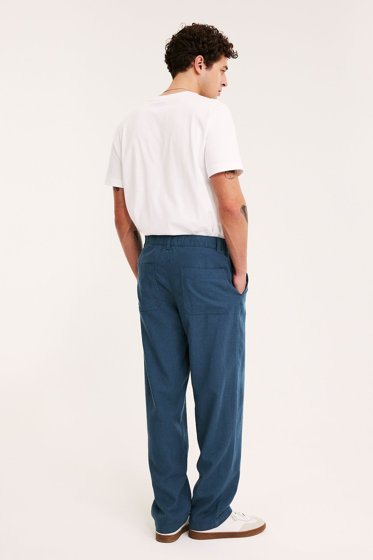 Mężczyzna z tyłu, z boku: niebieskie lniane spodnie o regularnym kroju, biały T-shirt i białe trampki.