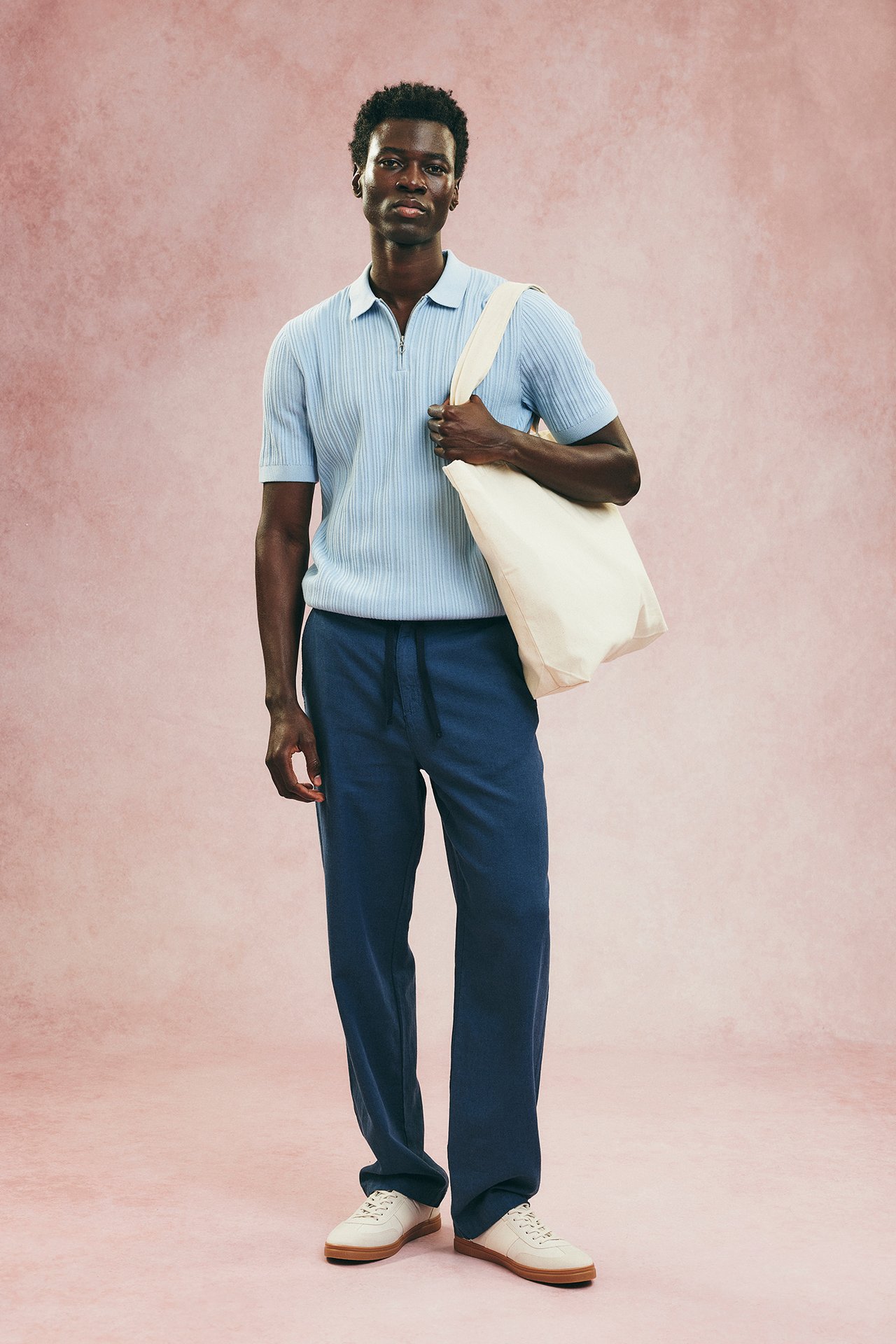 Etunäkymä: Mies sinisissä pellavasekoitehousuissa, vaaleansininen ribattu pikeepaita, valkoiset tennarit, pitää valkoista kangaskassia.