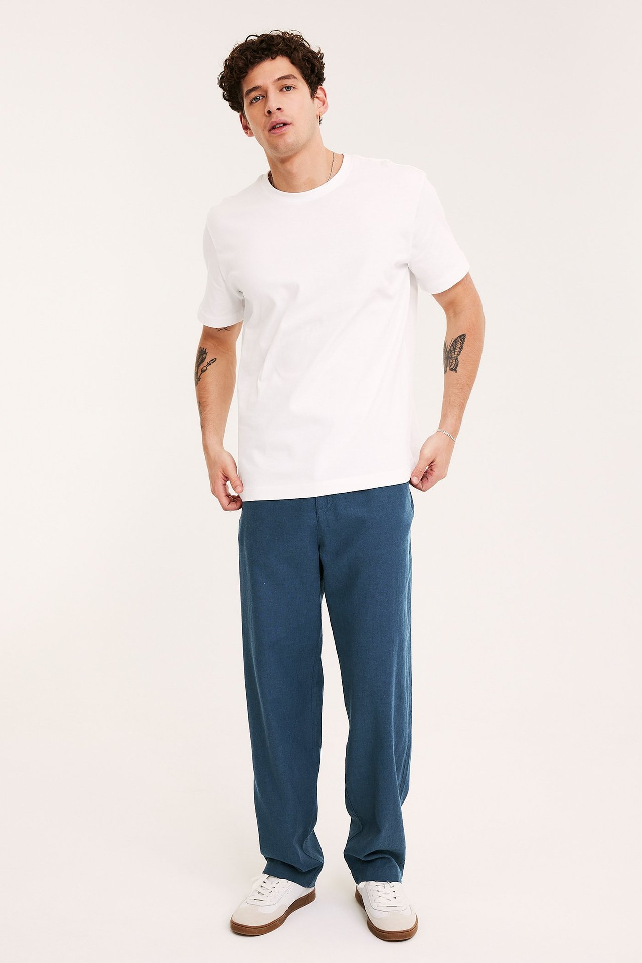 Widok z przodu mężczyzny w niebieskich spodniach z lnu, białej koszulce i białych tenisówkach.